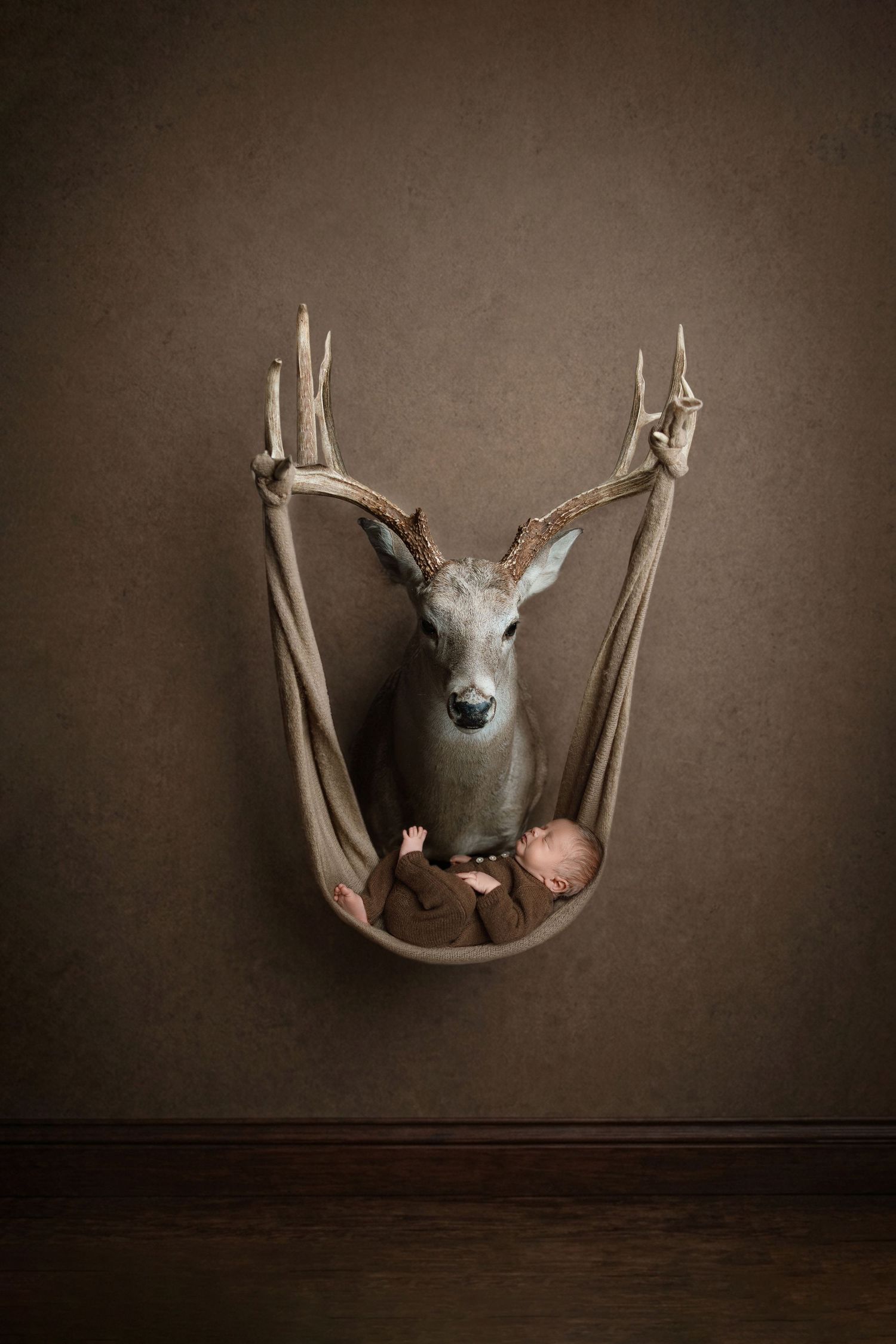 A newborn baby sleeps in a hammock made from antlers of a mounted deer head on a brown wall.
