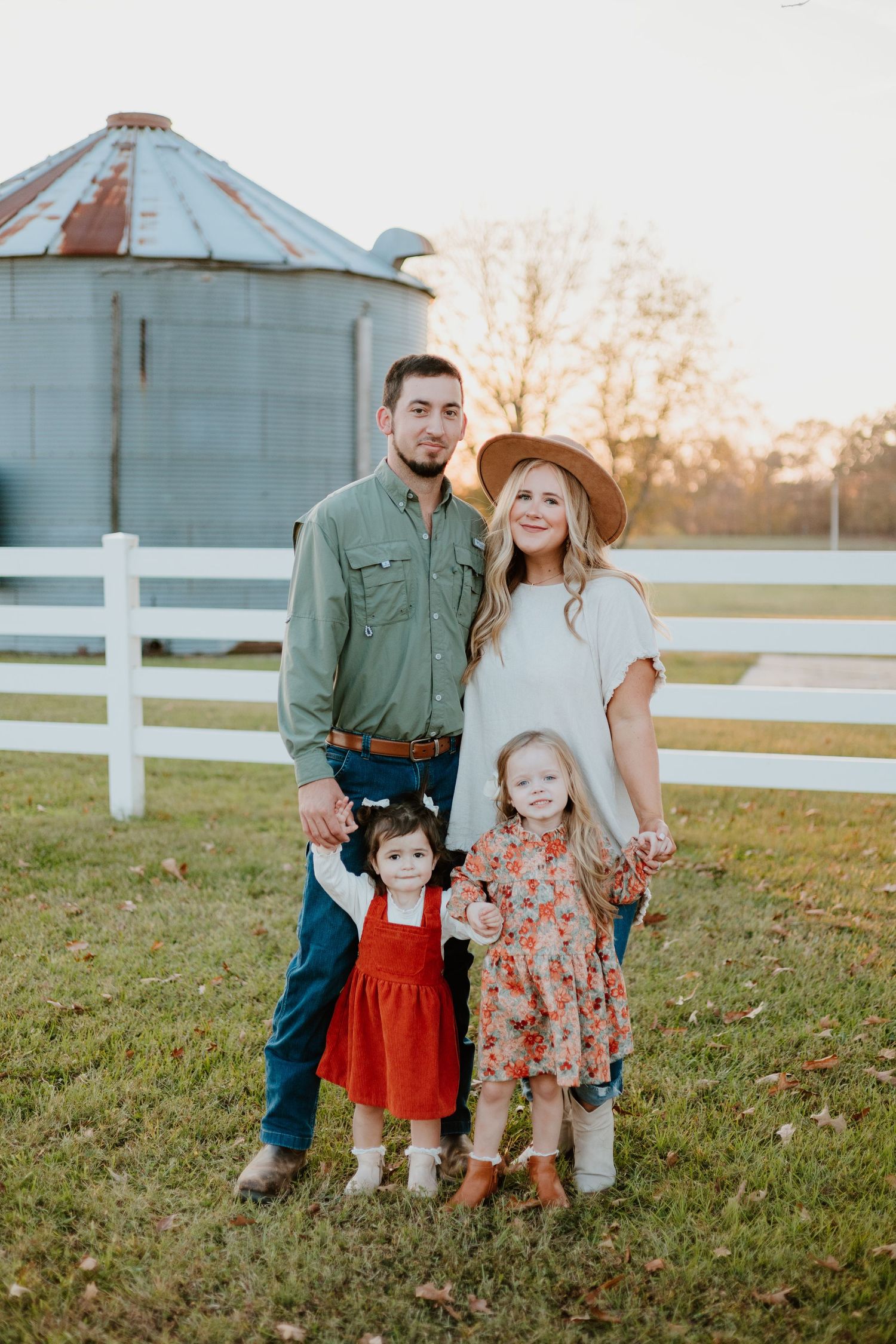 Family poses together in front of white fence and grain silo during sunset in rustic countryside setting.