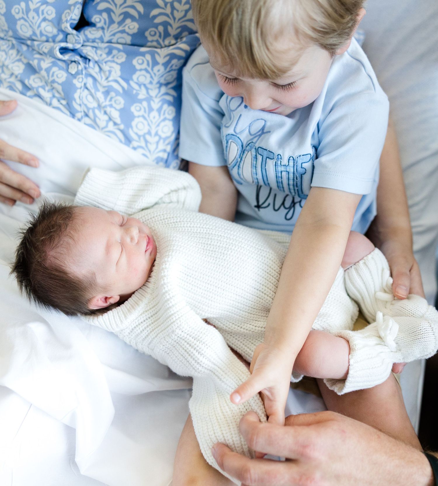 Toddler boy gently holds newborn baby sibling wrapped in white knit blanket on bed.