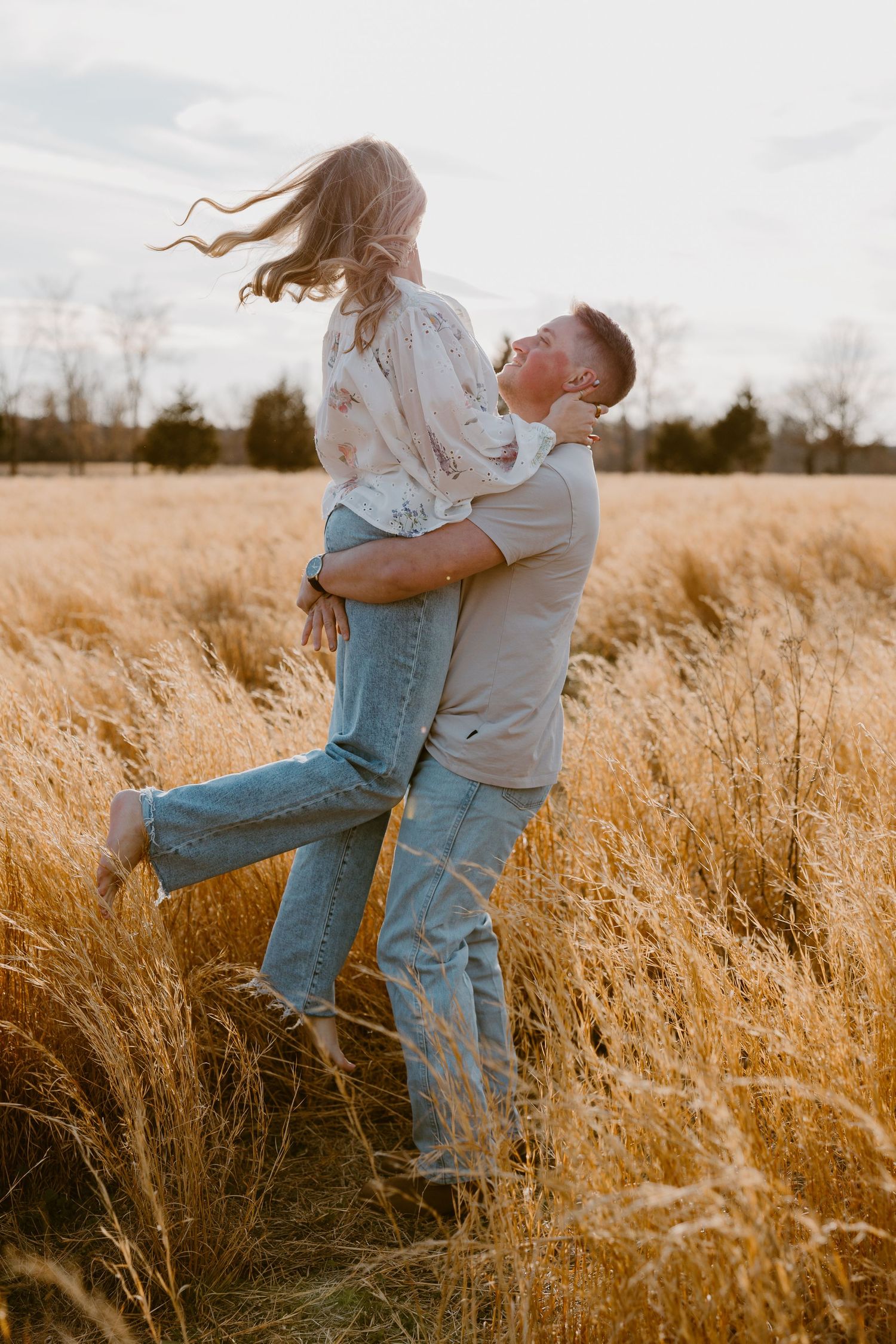 Romantic couple embraces in golden wheat field at sunset, with her hair flowing in the breeze.