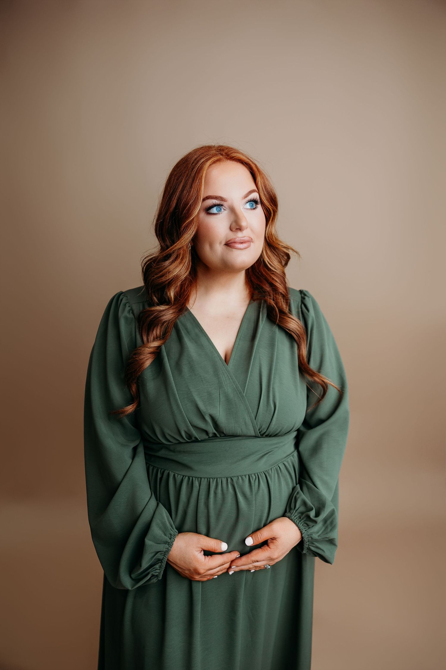 Woman wearing a long sleeve emerald green wrap dress poses against beige backdrop while holding phone.