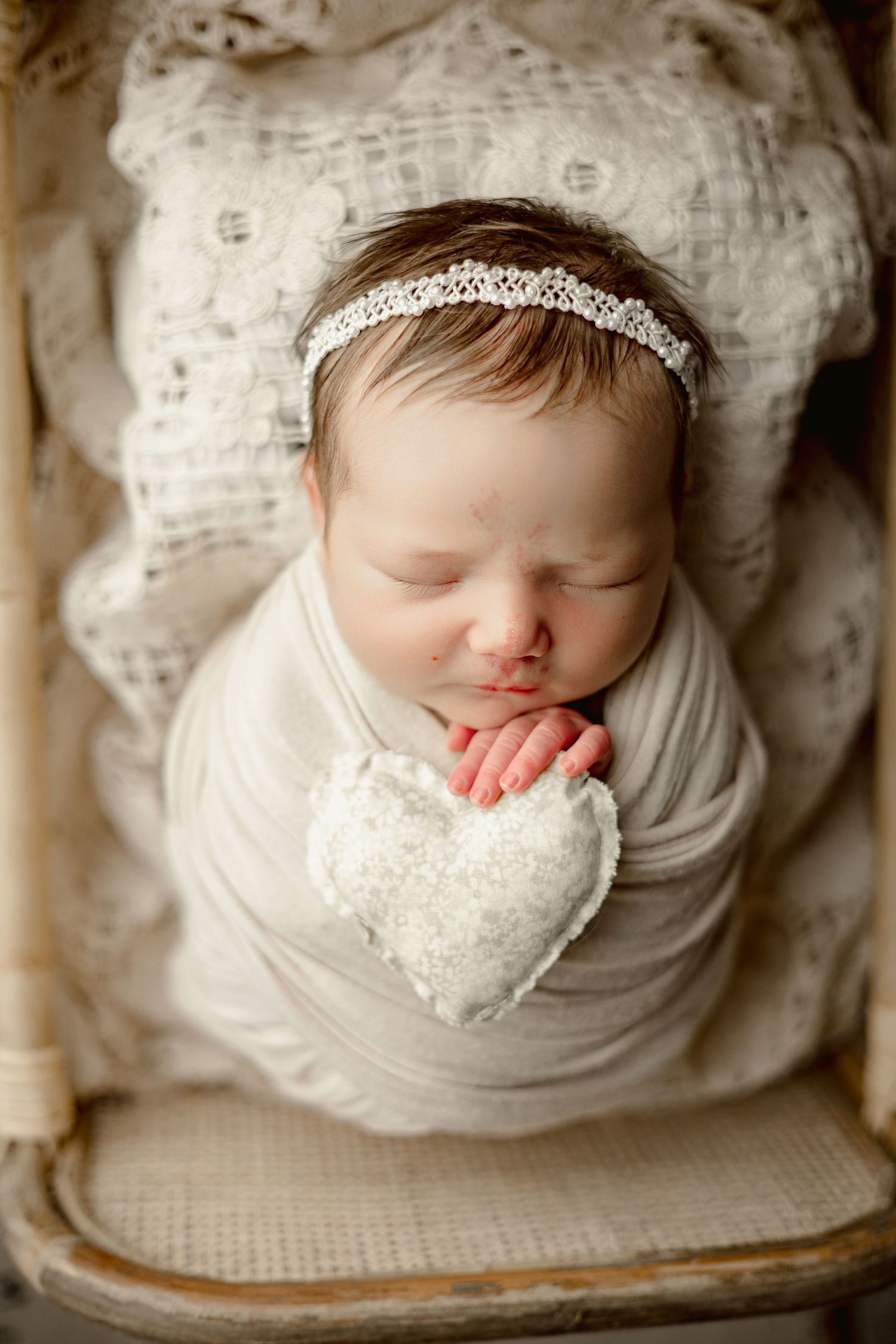 Sleeping newborn wrapped in white fabric with sparkly headband holds heart pillow while resting on vintage textured surface.