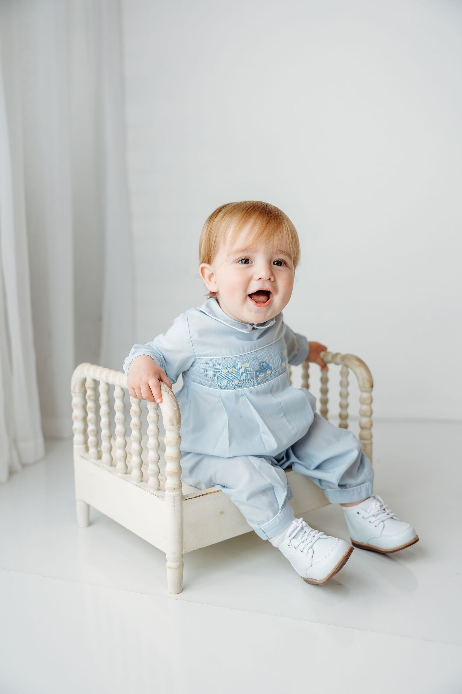 Baby wearing light blue outfit sits happily on antique white wooden spool bed against neutral background.