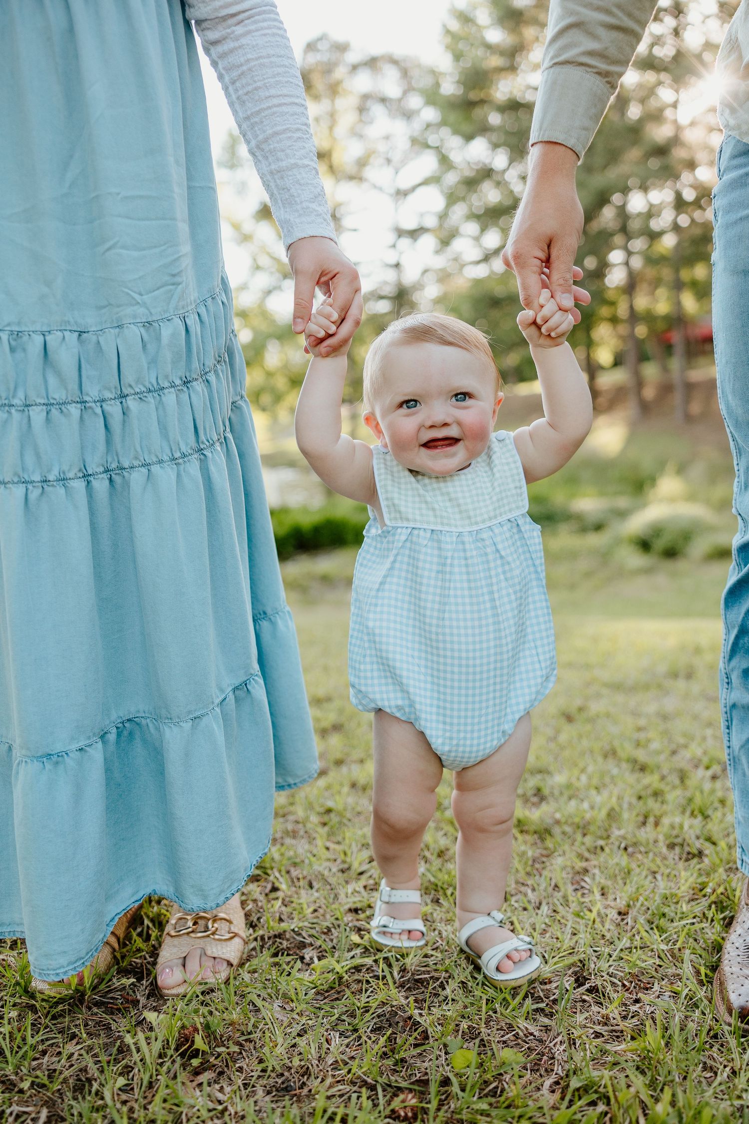Happy baby in blue gingham outfit holds hands with adults while standing on grass during golden hour.