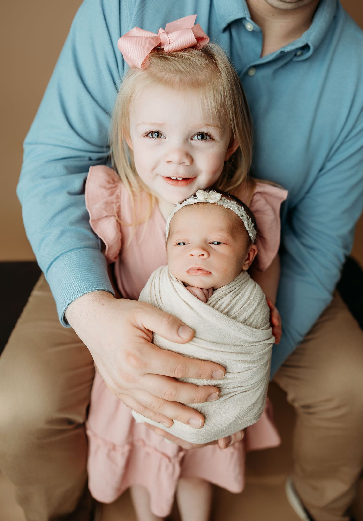 A baby wearing a cream swaddle and pink bow is held by someone in a turquoise shirt.