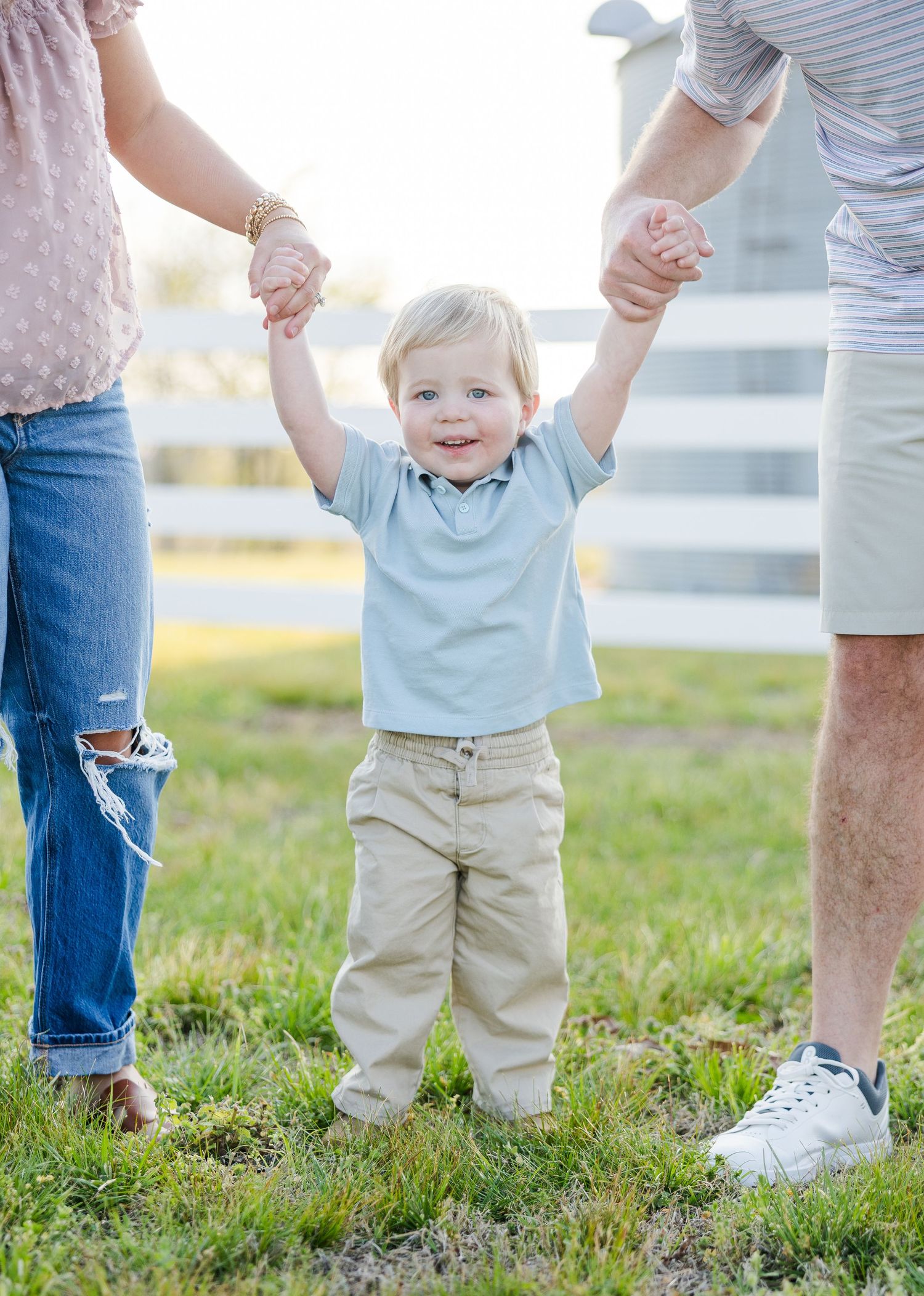 Happy toddler boy holding hands with parents while standing on green grass outdoors.