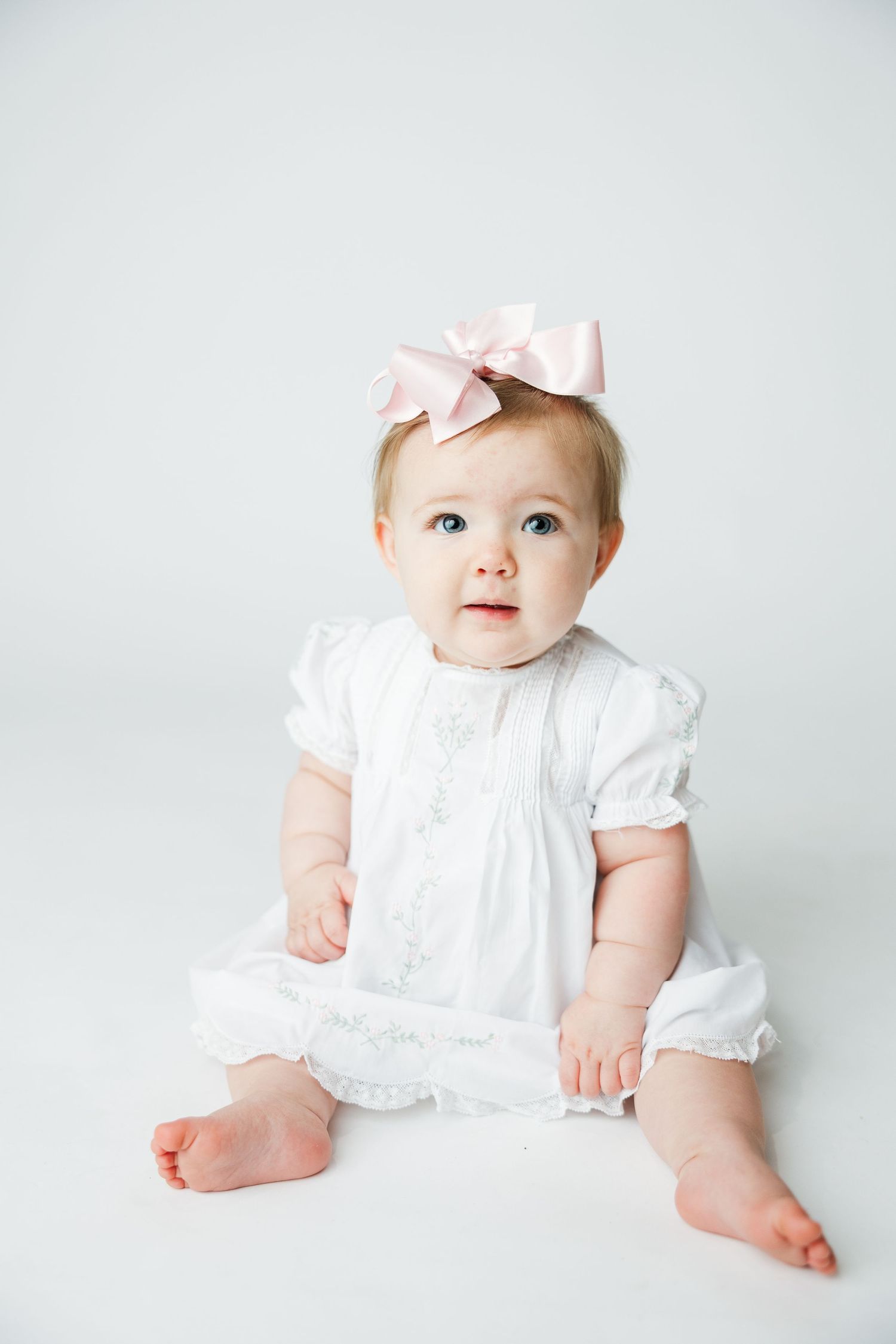 Sweet baby in white cotton dress with pink bow headband sitting on light background.