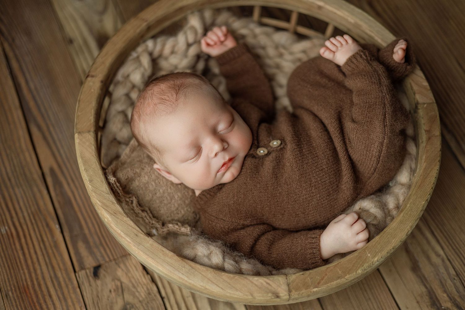 Sleeping newborn baby in brown knit outfit curled in rustic wooden basket with soft fur lining on wood floor.