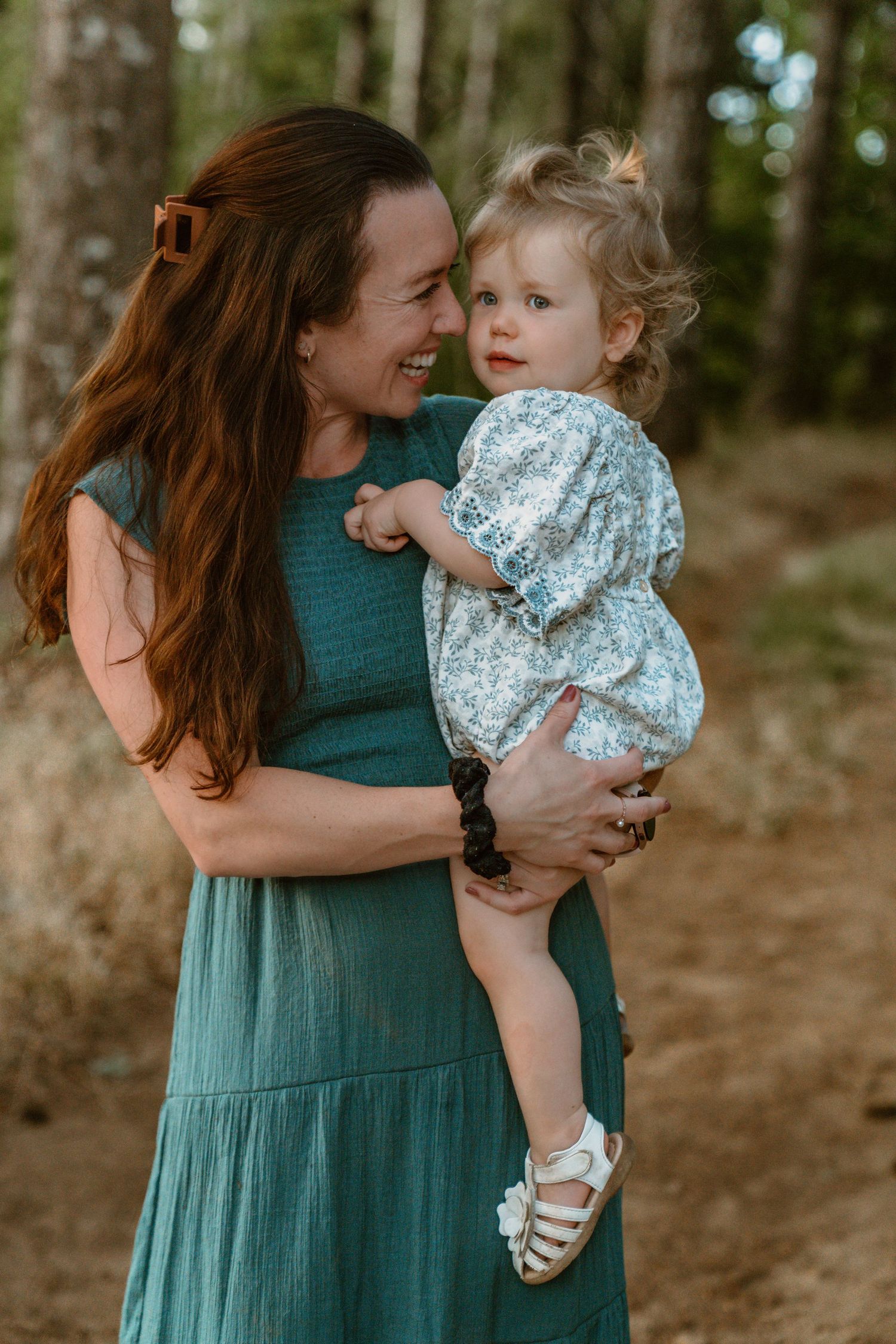 Mother in teal dress holding and laughing with toddler in floral outfit.