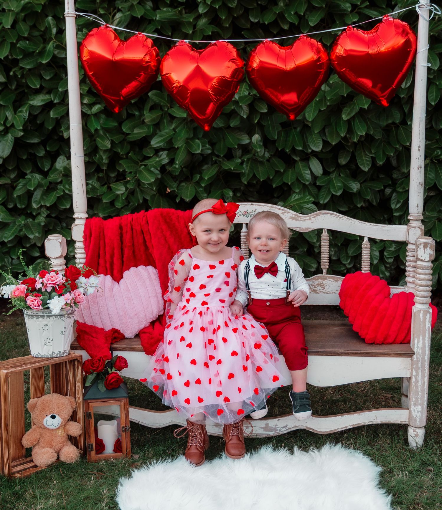 Two young children pose on a vintage bench with red heart balloons, Valentine's Day decor, and a teddy bear outdoors.