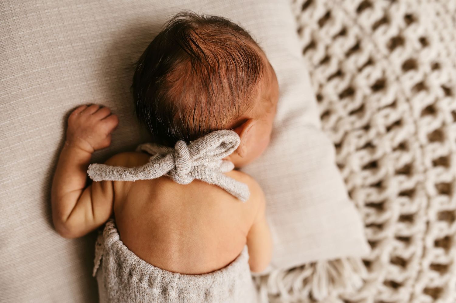 Newborn baby lying on a knit blanket with a bow, photographed from above in soft neutral tones.