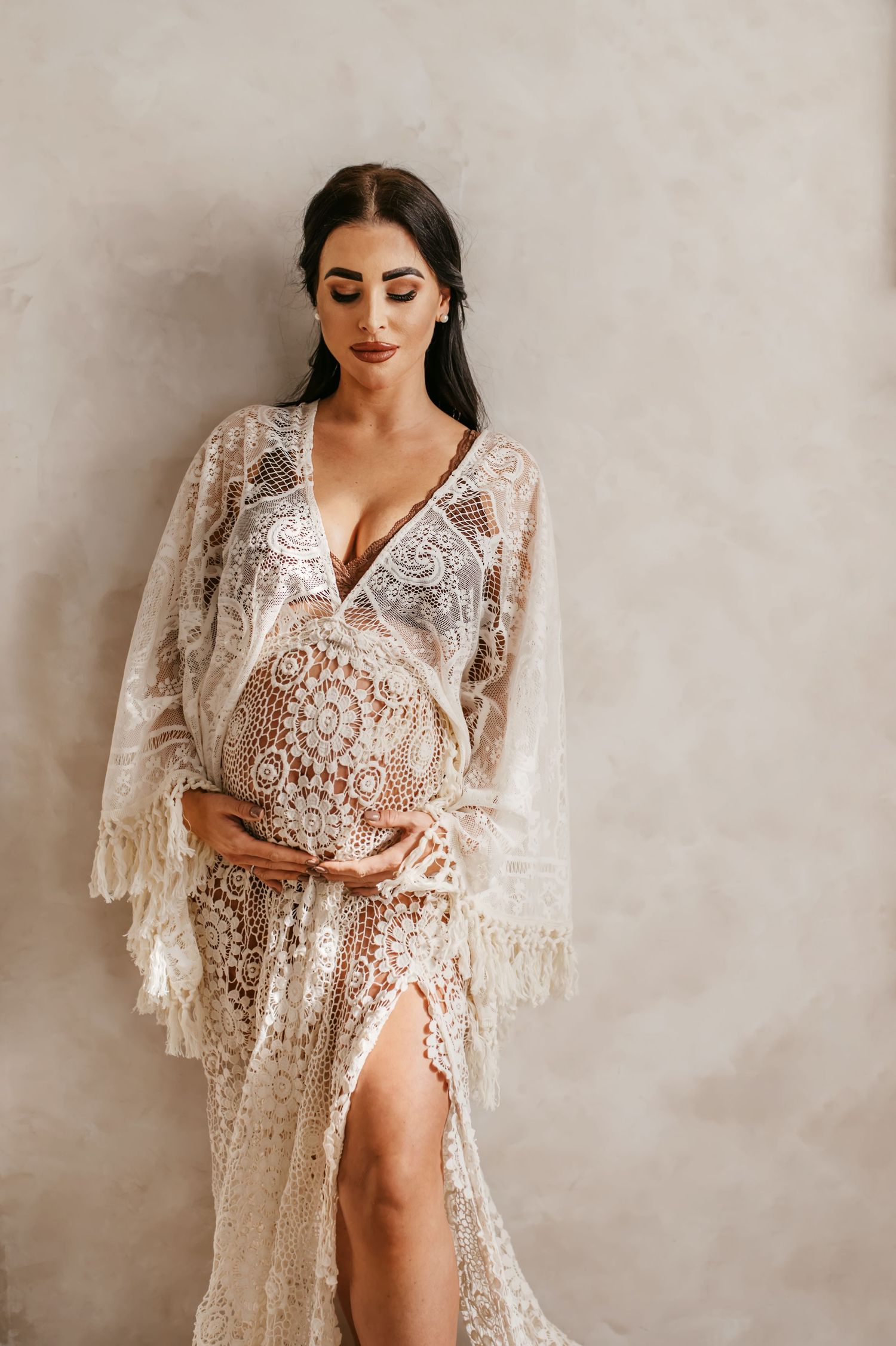 Elegant white lace maternity dress with bell sleeves and deep side slit against neutral backdrop.