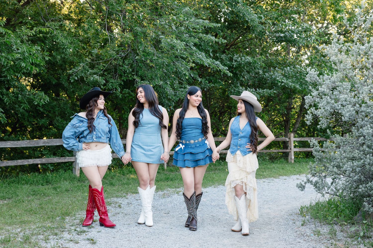 Four women in coordinated denim outfits and cowboy boots walk together on a gravel path surrounded by lush green trees.