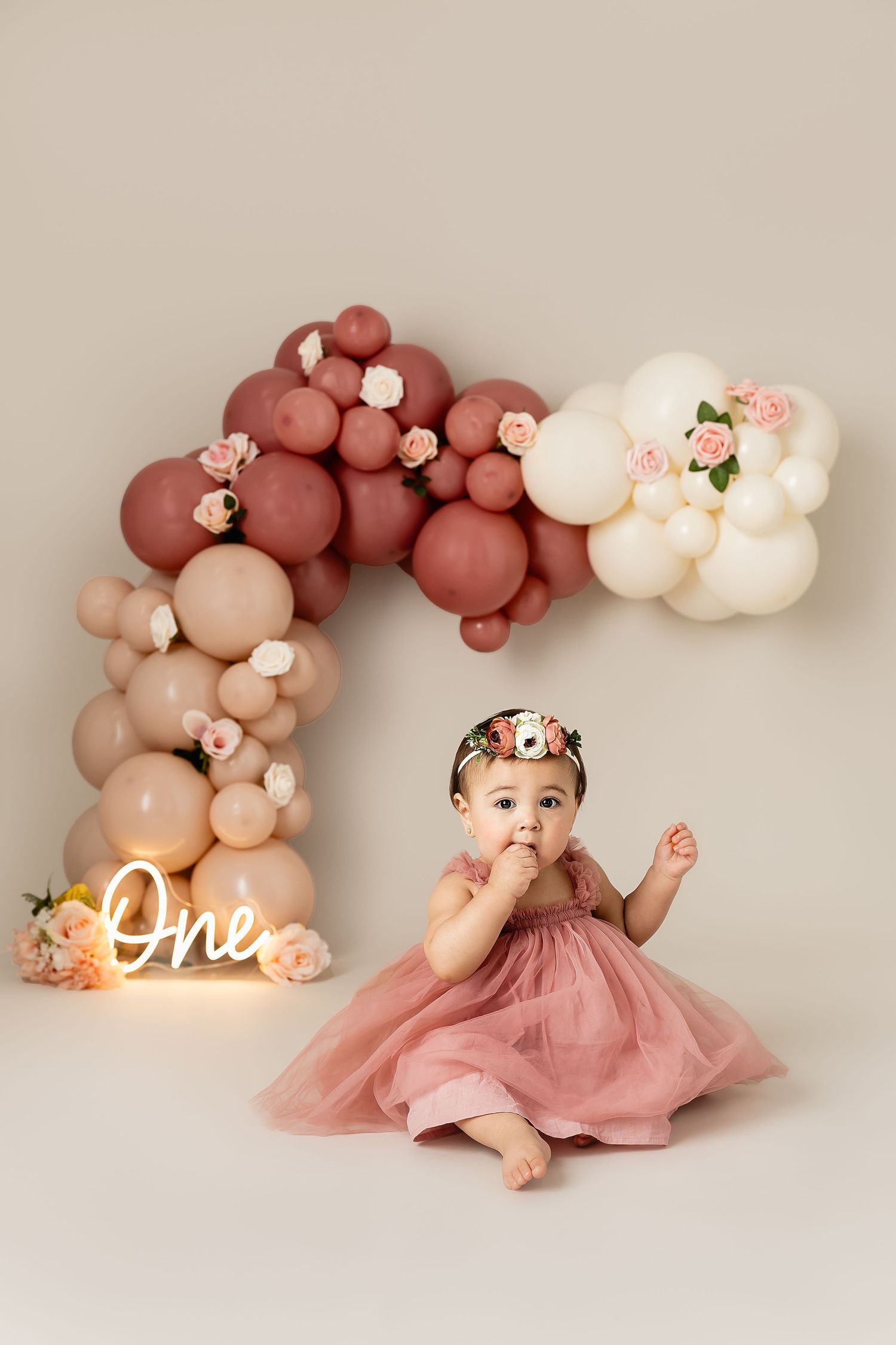 First birthday celebration setup with dusty rose balloon arch, neon 'One' sign, and floral decorations against neutral backdrop.