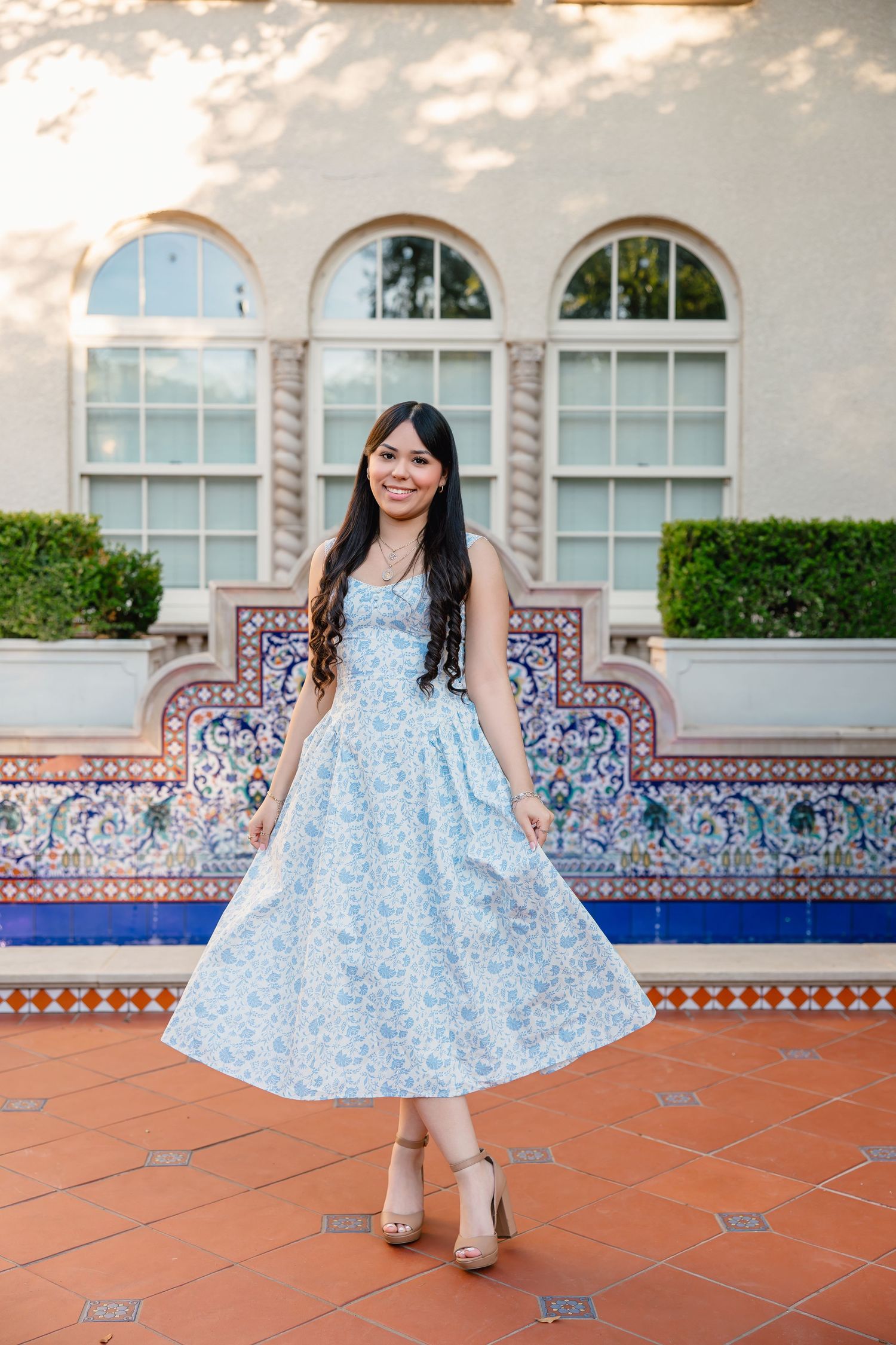 Young woman in a light blue floral midi dress twirling on a terracotta patio with decorative tile bench backdrop.