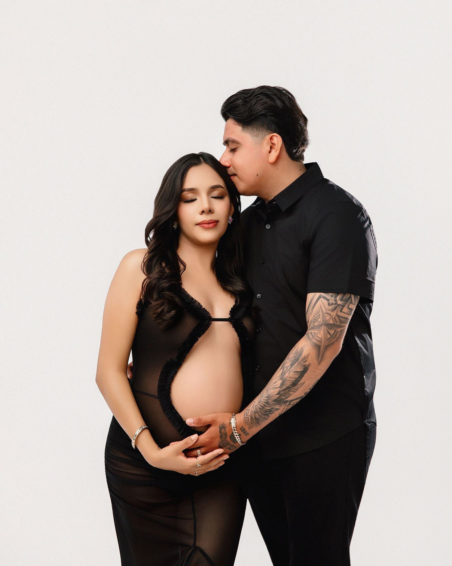 Couple in black outfits embraces intimately during maternity photo shoot against white backdrop.