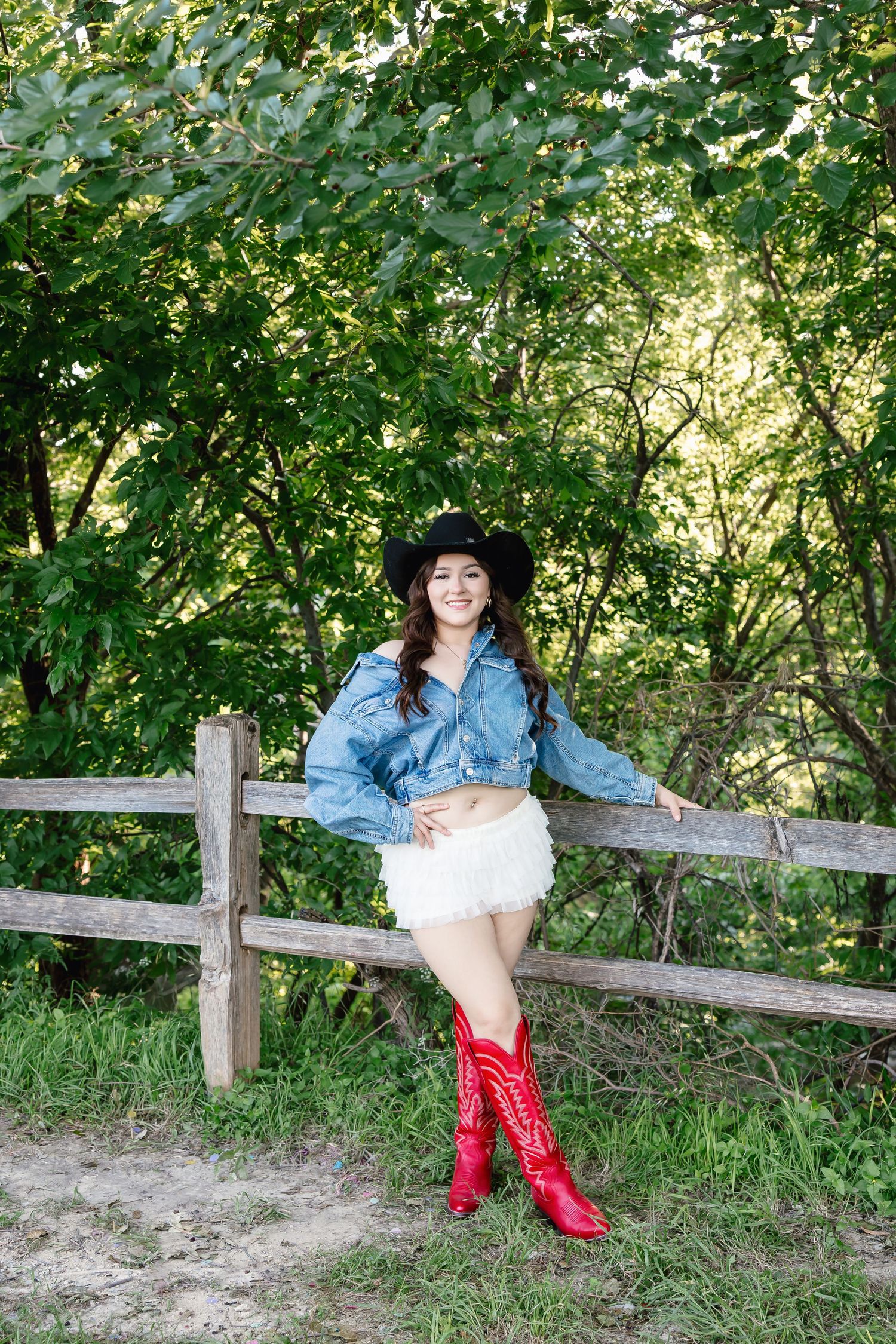 Young woman in denim jacket, white skirt, red cowboy boots and black hat leaning on wooden fence outdoors.