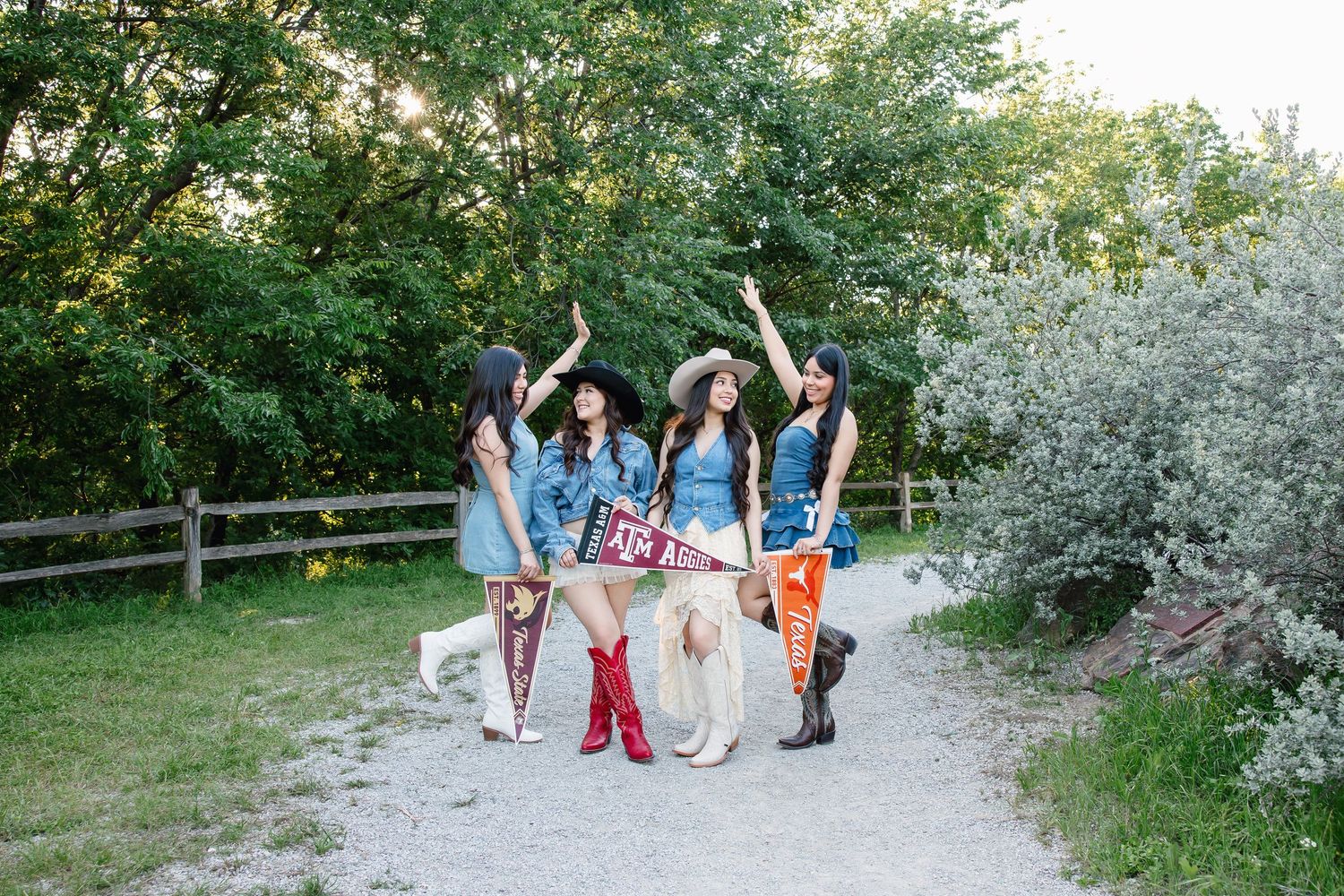 Four women in cowgirl outfits with boots and hats pose on a gravel path surrounded by lush green trees.