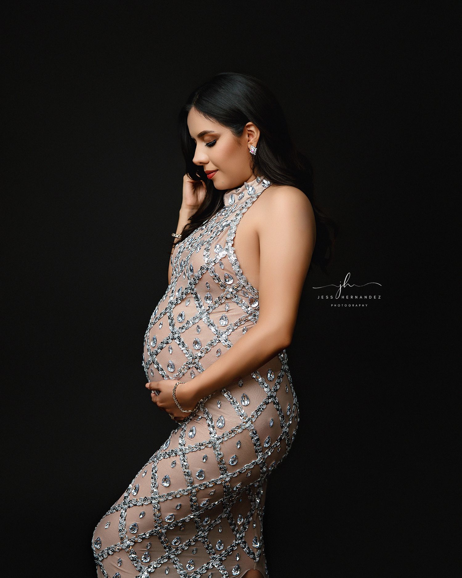 Elegant maternity photoshoot featuring a sheer beaded gown against a dark backdrop in dramatic studio lighting.
