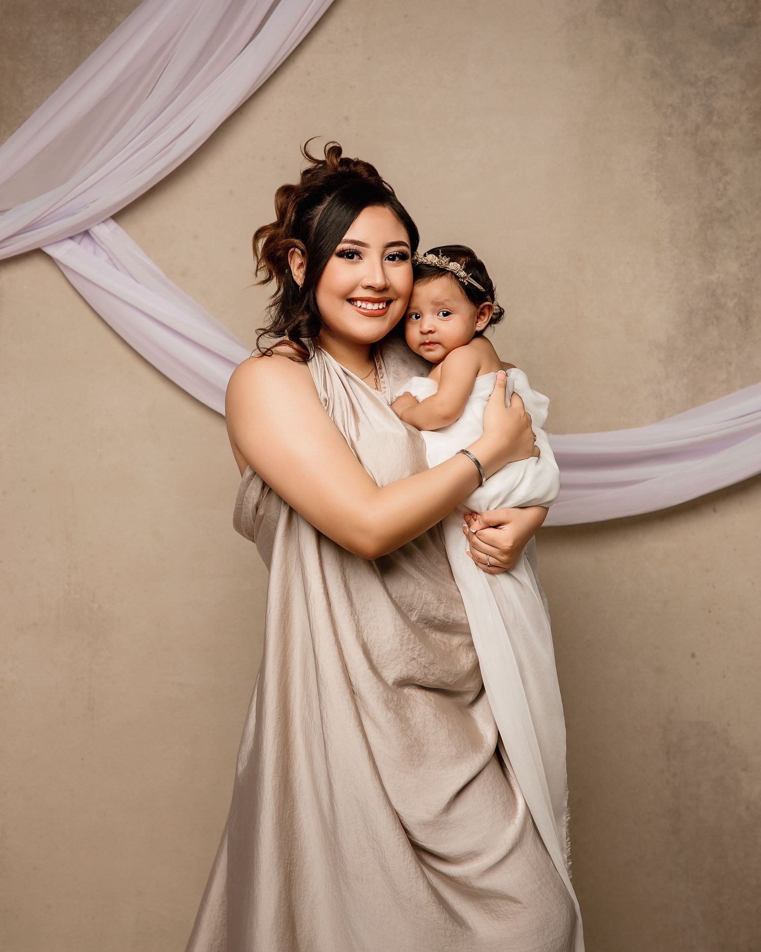 A mother and baby in matching cream dresses pose together against a textured wall with draped fabric backdrop.