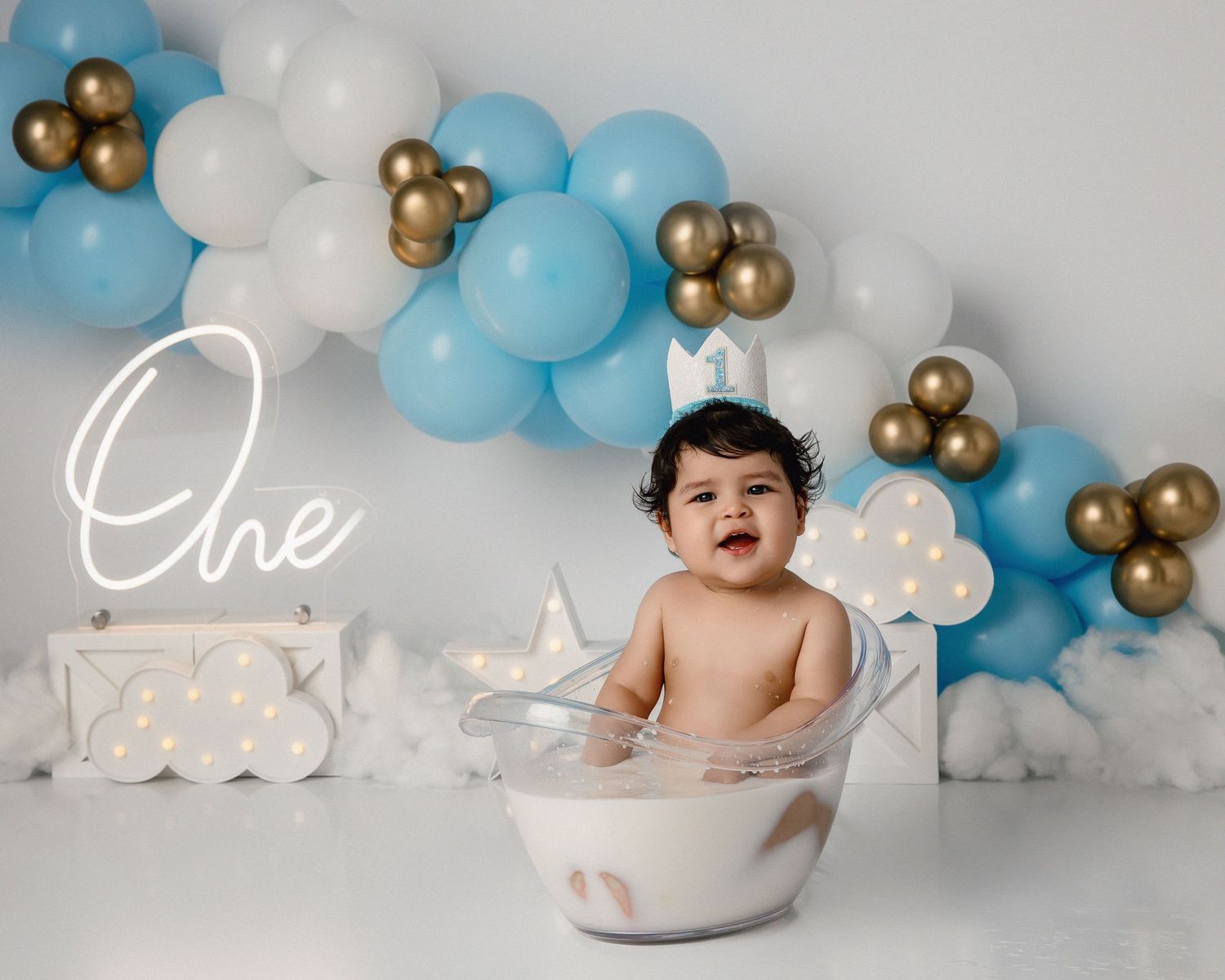 Baby's first birthday cake smash photo with blue, white, and gold balloons, sitting in a milk bath tub.