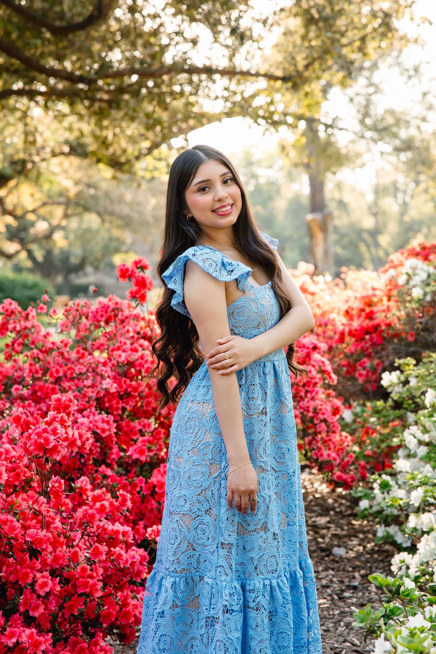 Young woman in blue lace maxi dress smiling among vibrant red azalea bushes in a sunlit garden.