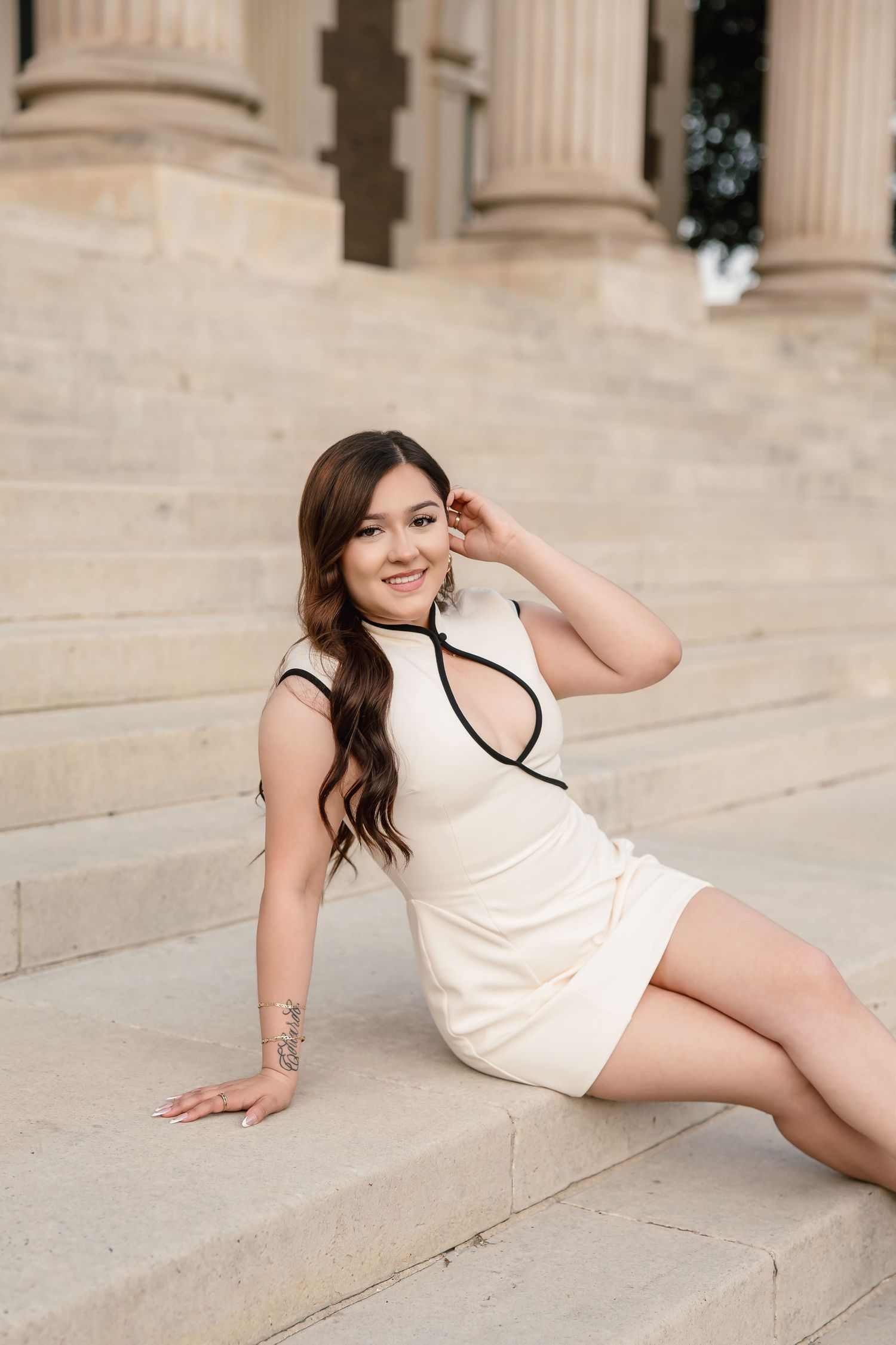 Young woman in white mini dress with black trim sits gracefully on stone steps near classical columns.