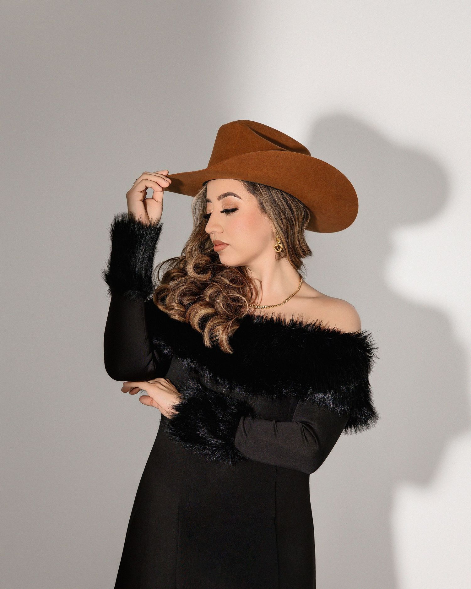 Model in brown cowboy hat and black fur-trimmed coat poses against white background in fashion photoshoot series.