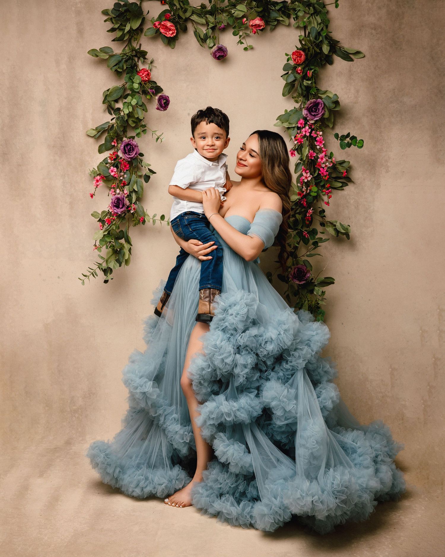 Mother and child in matching blue tulle dresses pose against floral backdrop in studio portrait session.
