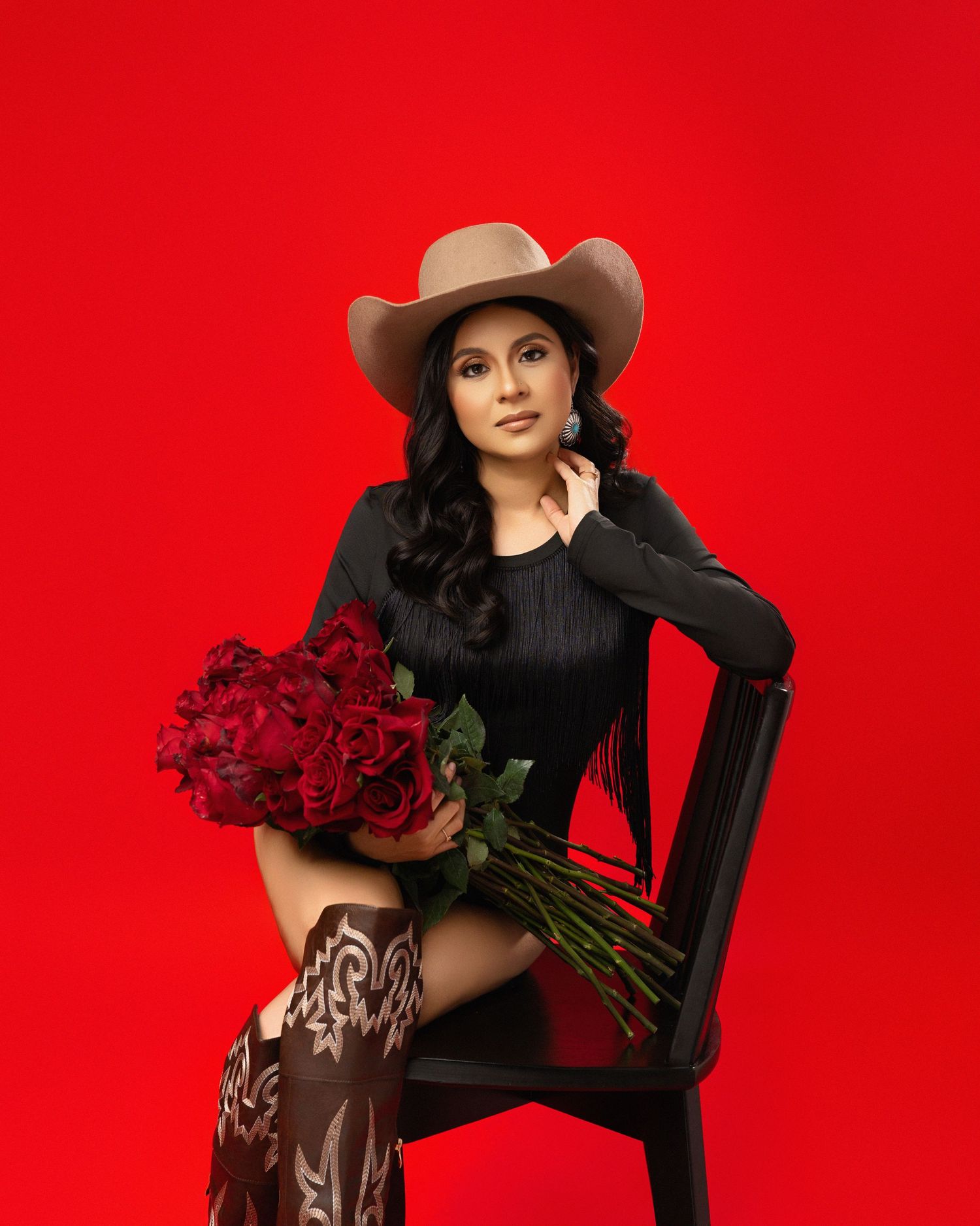 Western-inspired fashion portrait with cowboy hat and boots against vibrant red backdrop, holding red roses.