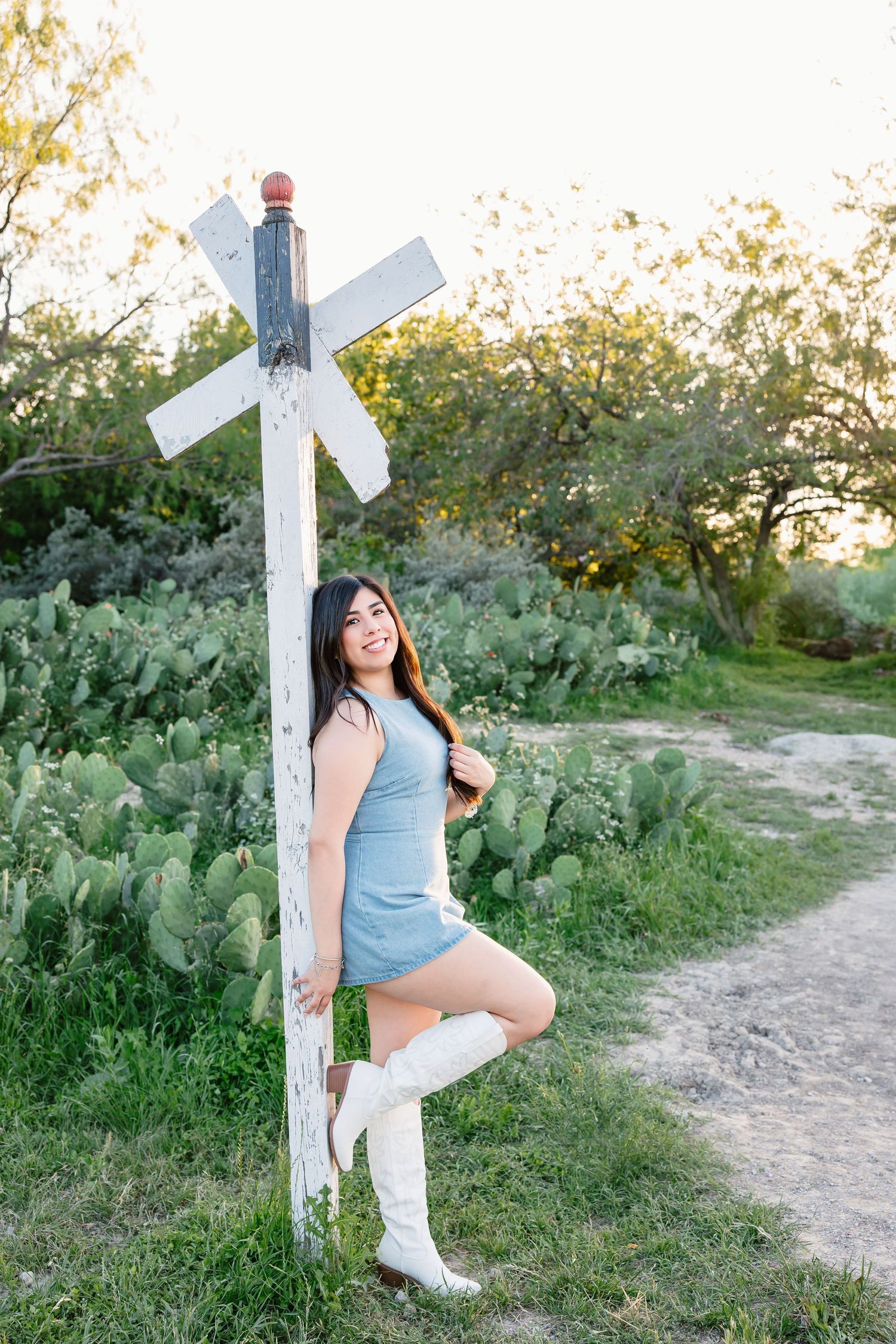 Young woman in blue dress and white cowboy boots poses beside a rustic railroad crossing sign amid cacti.