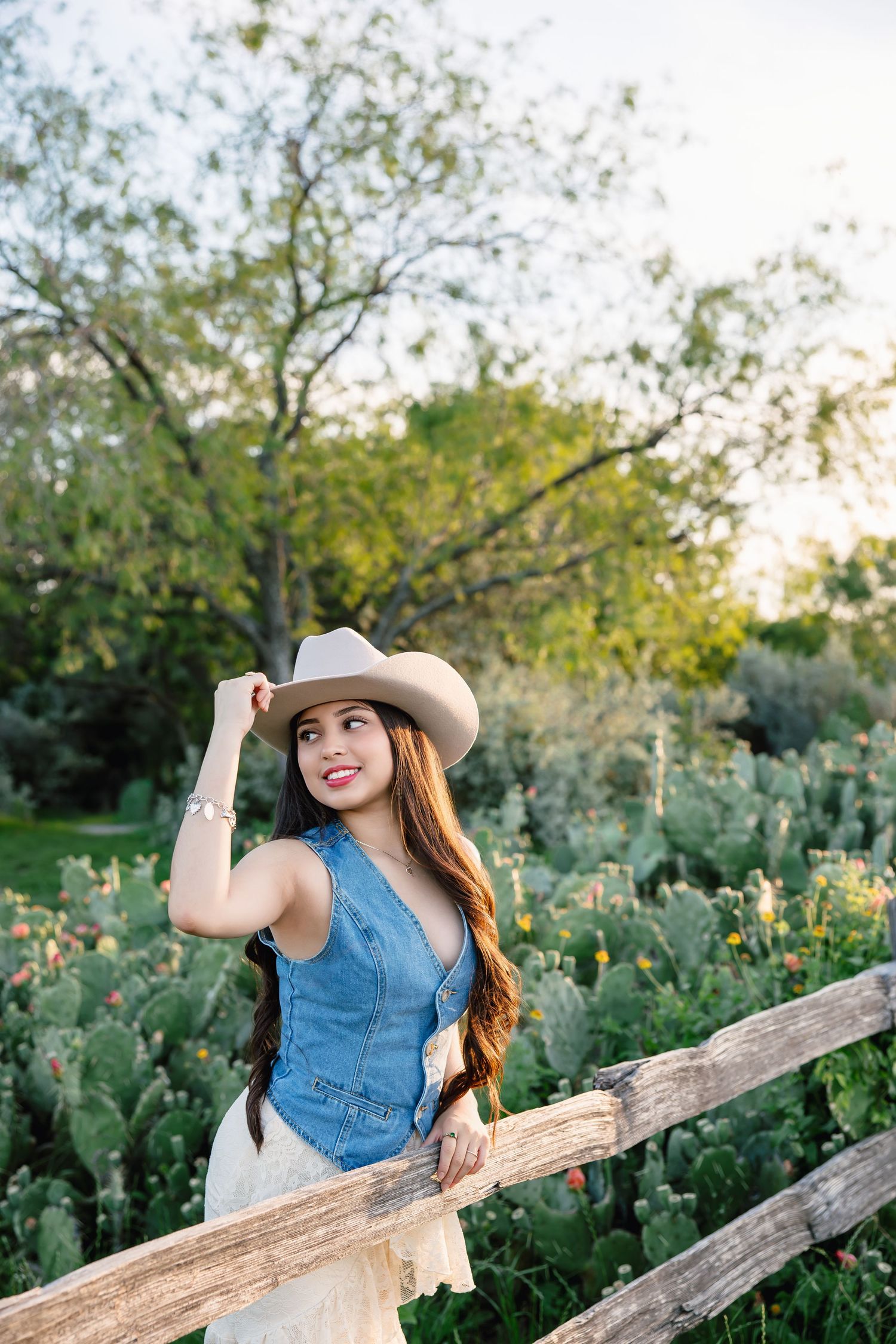 Young woman in cowboy hat and denim vest leaning on rustic wooden fence with cactus garden backdrop.