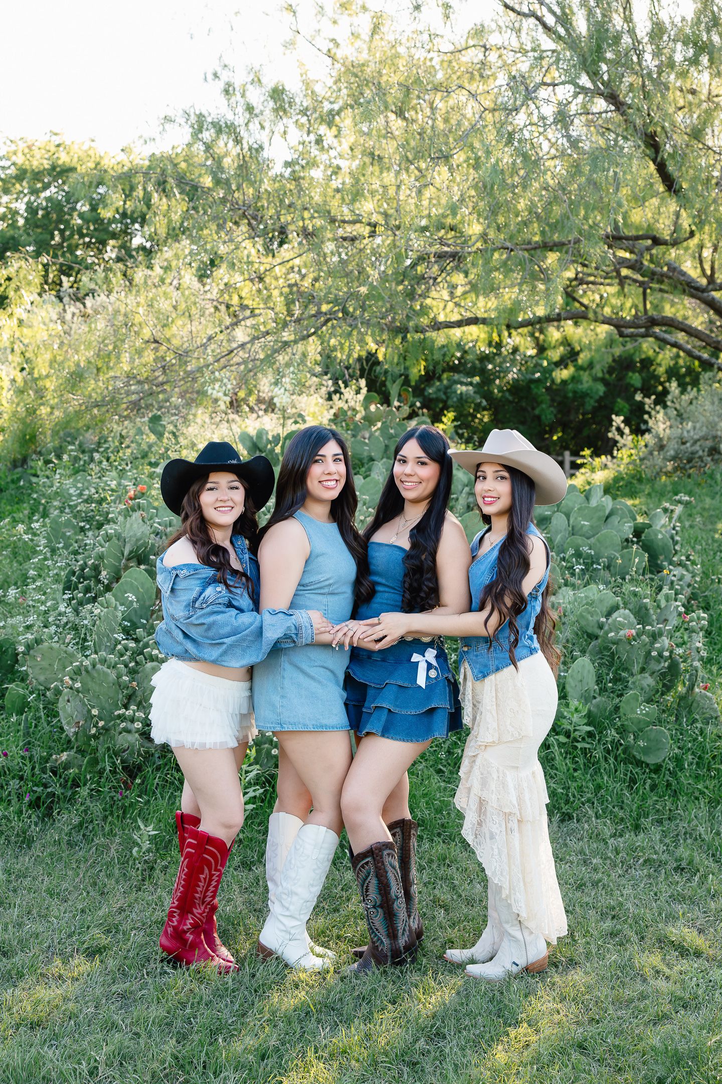 Four young women in western cowboy hats, denim outfits, and boots pose together outdoors in a green natural setting.