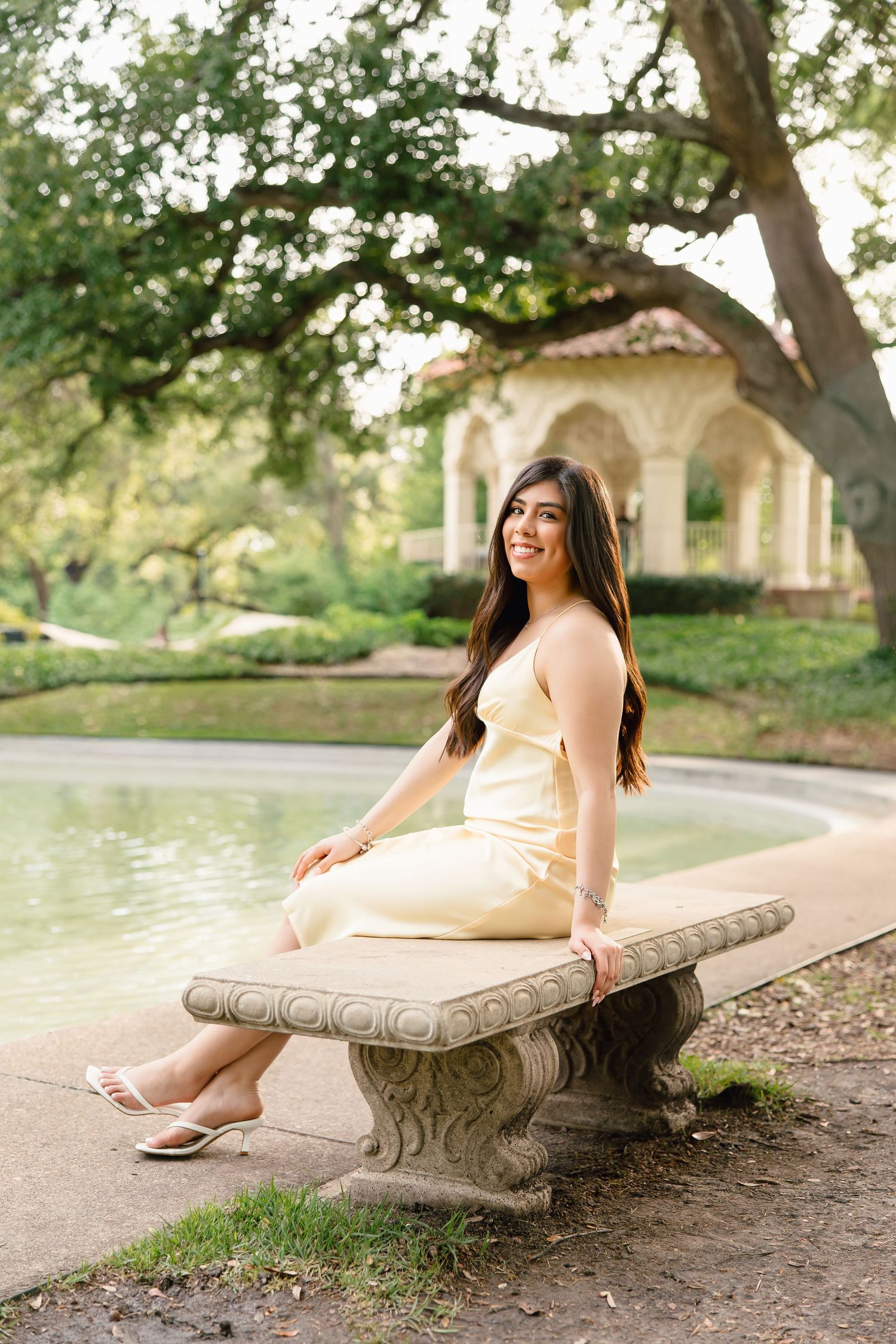 Young woman in yellow dress sitting on stone bench by garden pond with arched pavilion backdrop.
