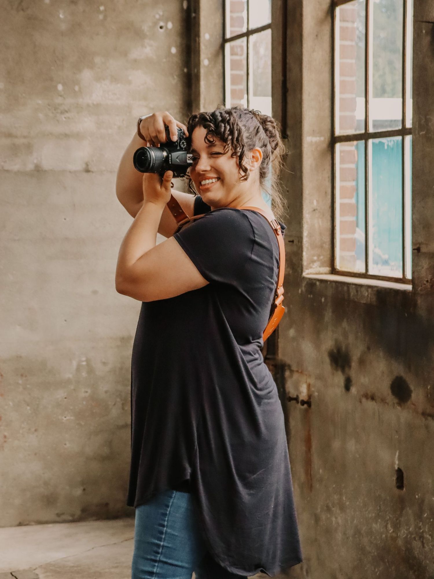 Een fotograaf in een casual zwarte jurk en spijkerbroek houdt een professionele camera vast bij rustieke ramen in een vintage gebouw.