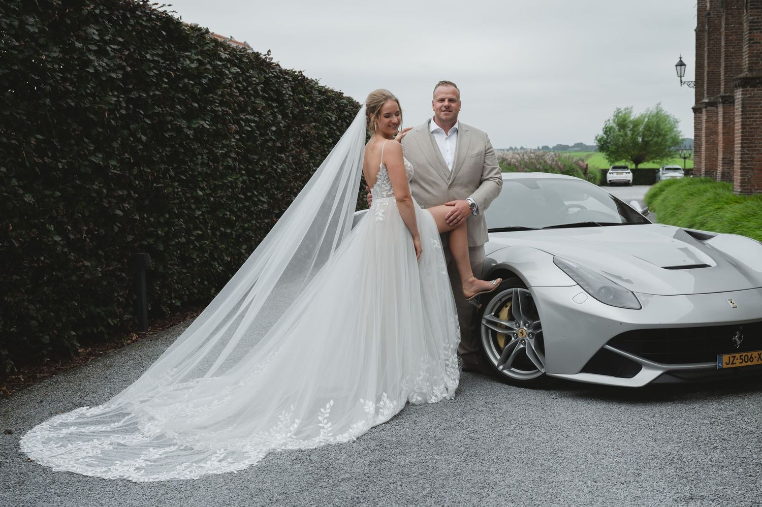 Een trouwpaar poseert naast een zilveren Ferrari sportwagen, waarbij de bruid een vloeiende witte jurk en sluier draagt.
