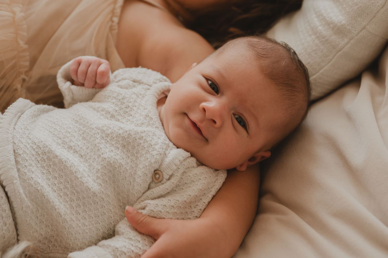 Baby in een witte romper met stippen ligt tevreden op wit beddengoed in warm natuurlijk licht.