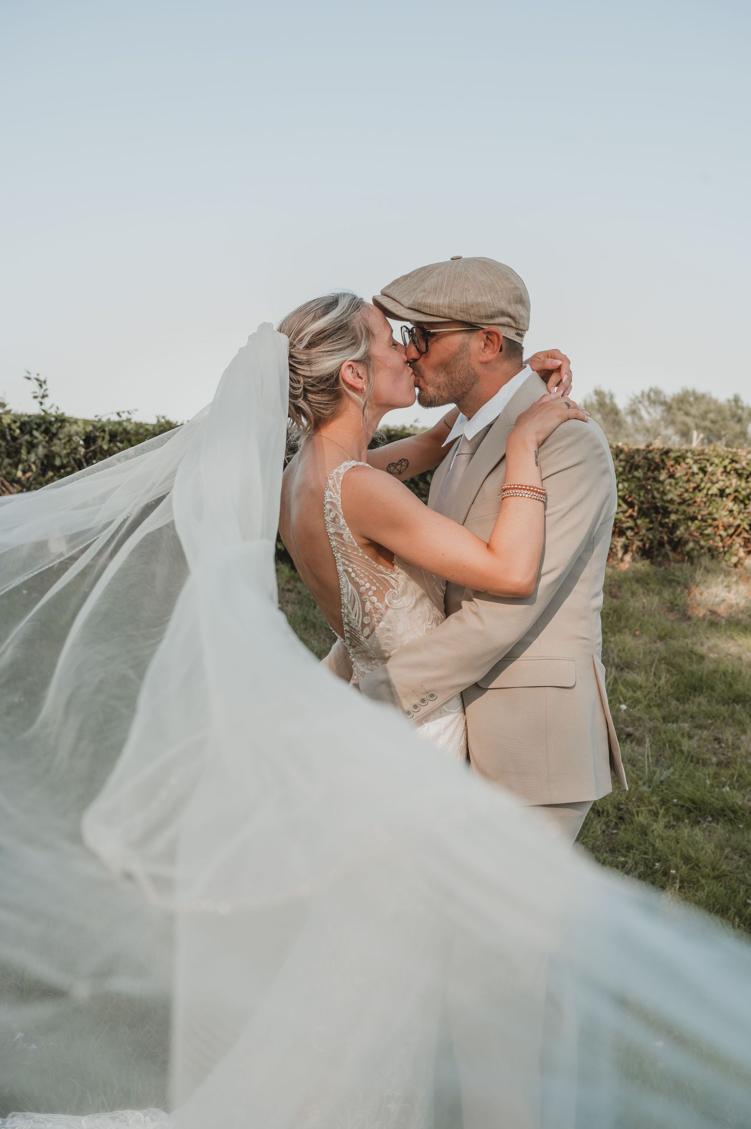 Een romantische trouwfoto met een wapperende witte sluier op de voorgrond en groene bladeren in een natuurlijke buitenomgeving.
