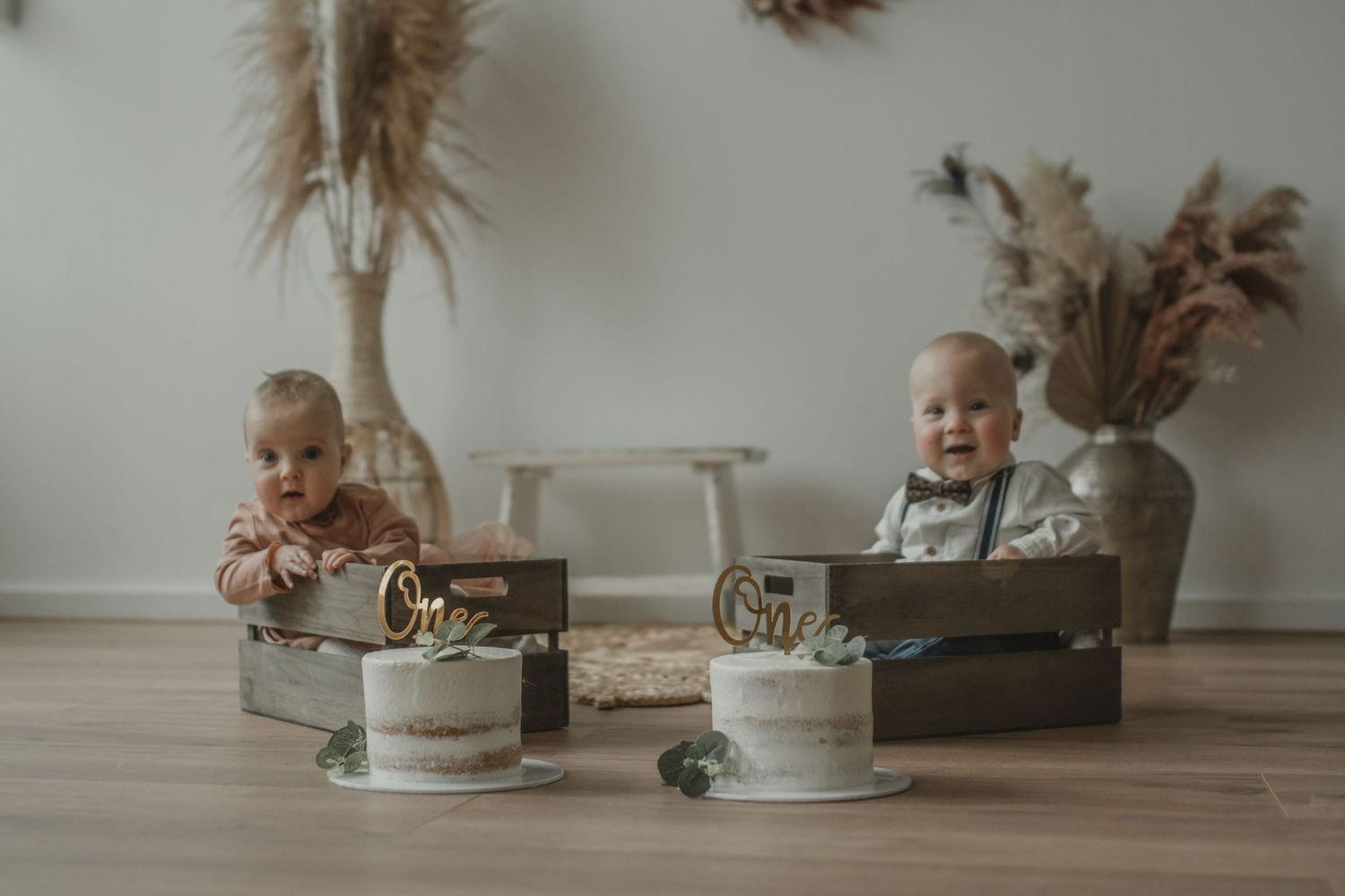 Twee kinderen zitten voor eenvoudige, matte verjaardagstaarten, versierd in gedempte tinten, in een minimalistische setting met pampasgras.