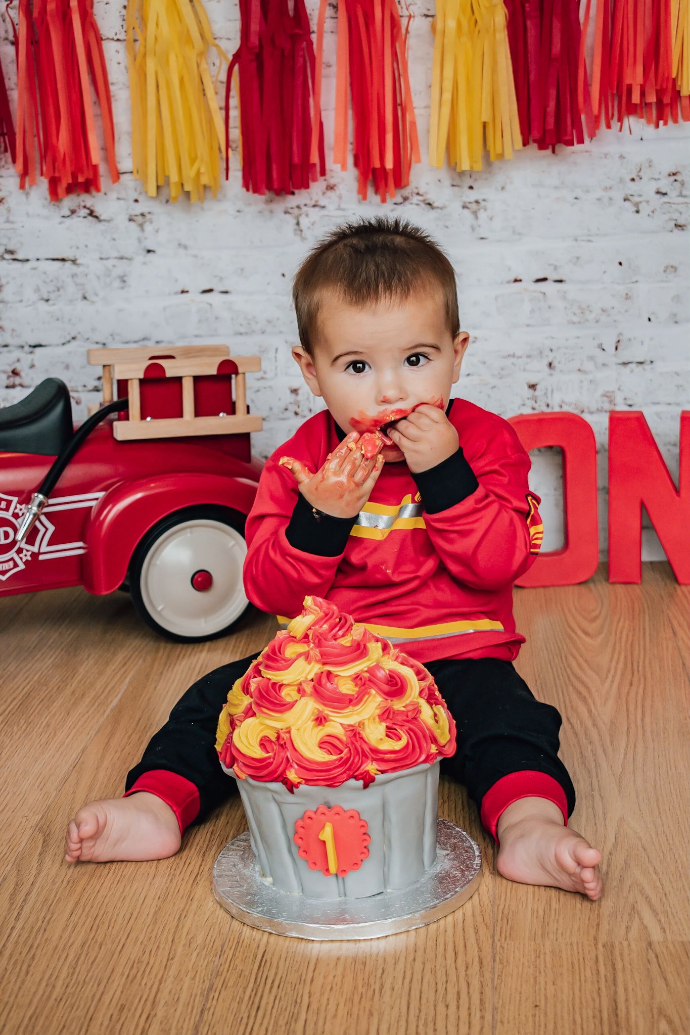 Een baby in het rood zit op een houten vloer met een grote smash cake en speelgoedwagentje, voor een feestelijk backdrop van crêpepapier.