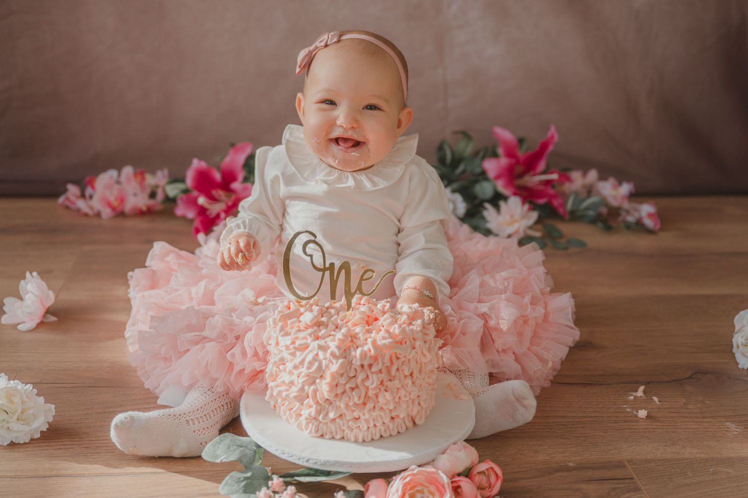 Een baby met een wit shirt met de tekst ‘One’ zit met een roze smash cake en een tule rokje te midden van bloemenversiering voor een eerste