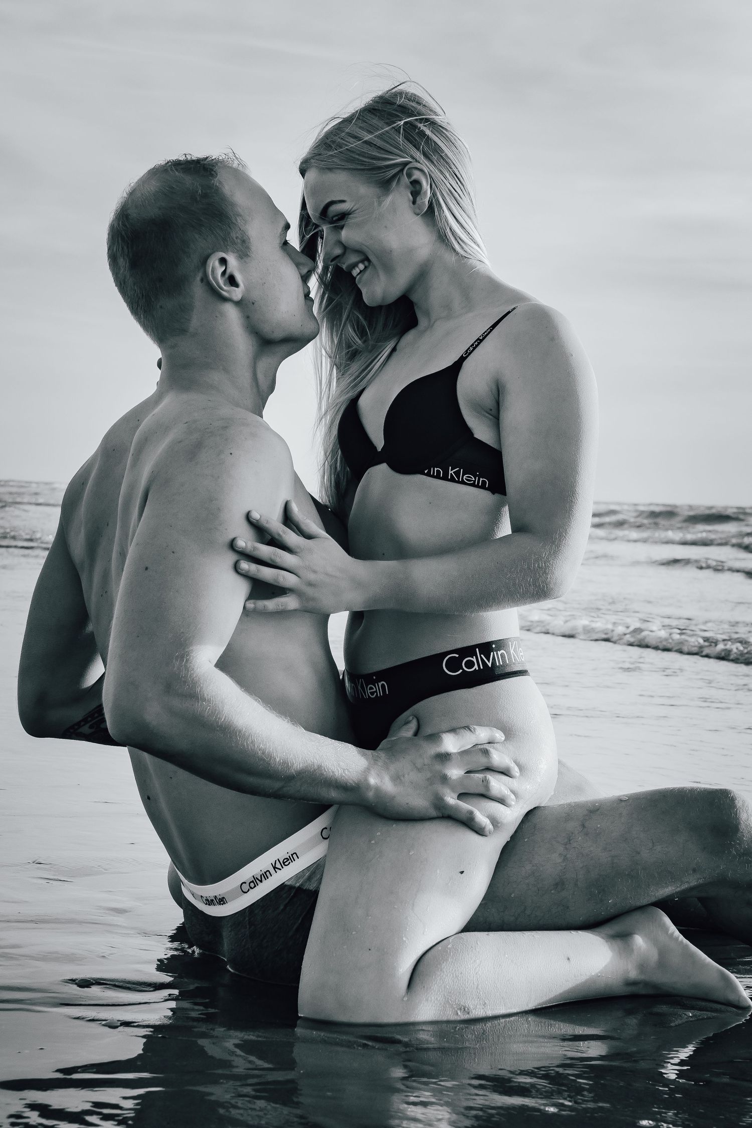 Intieme zwart-wit strandfoto van een stel in Calvin Klein ondergoed die elkaar omhelzen in ondiep oceaanwater.