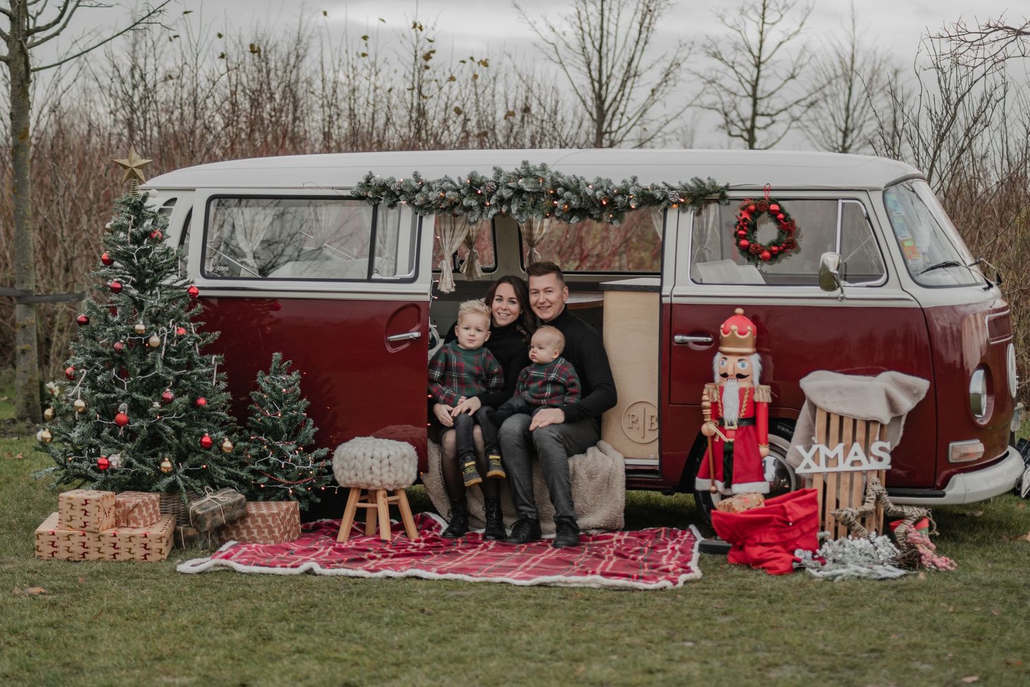 Een feestelijke kerstsfeer met een klassieke rood-witte VW-bus, een versierde kerstboom en geruite dekens op het gras.