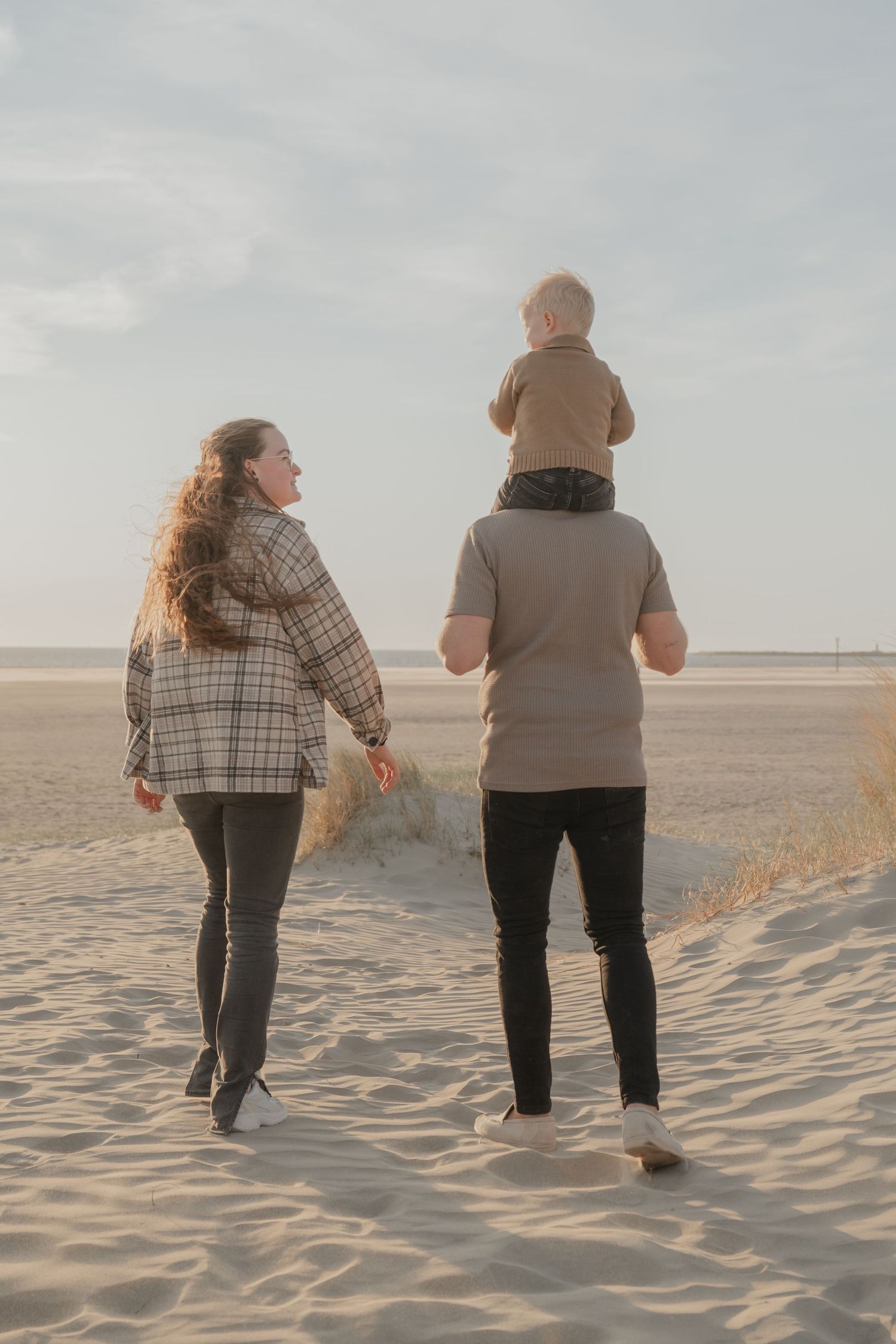 Een gezin wandelt samen over het zandstrand bij zonsondergang, met een klein kind op de schouders.