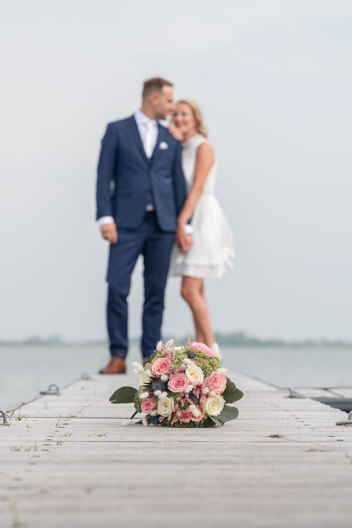 Bruidsboeket met roze en witte bloemen op een houten steiger, terwijl het bruidspaar vaag op de achtergrond bij het water staat.