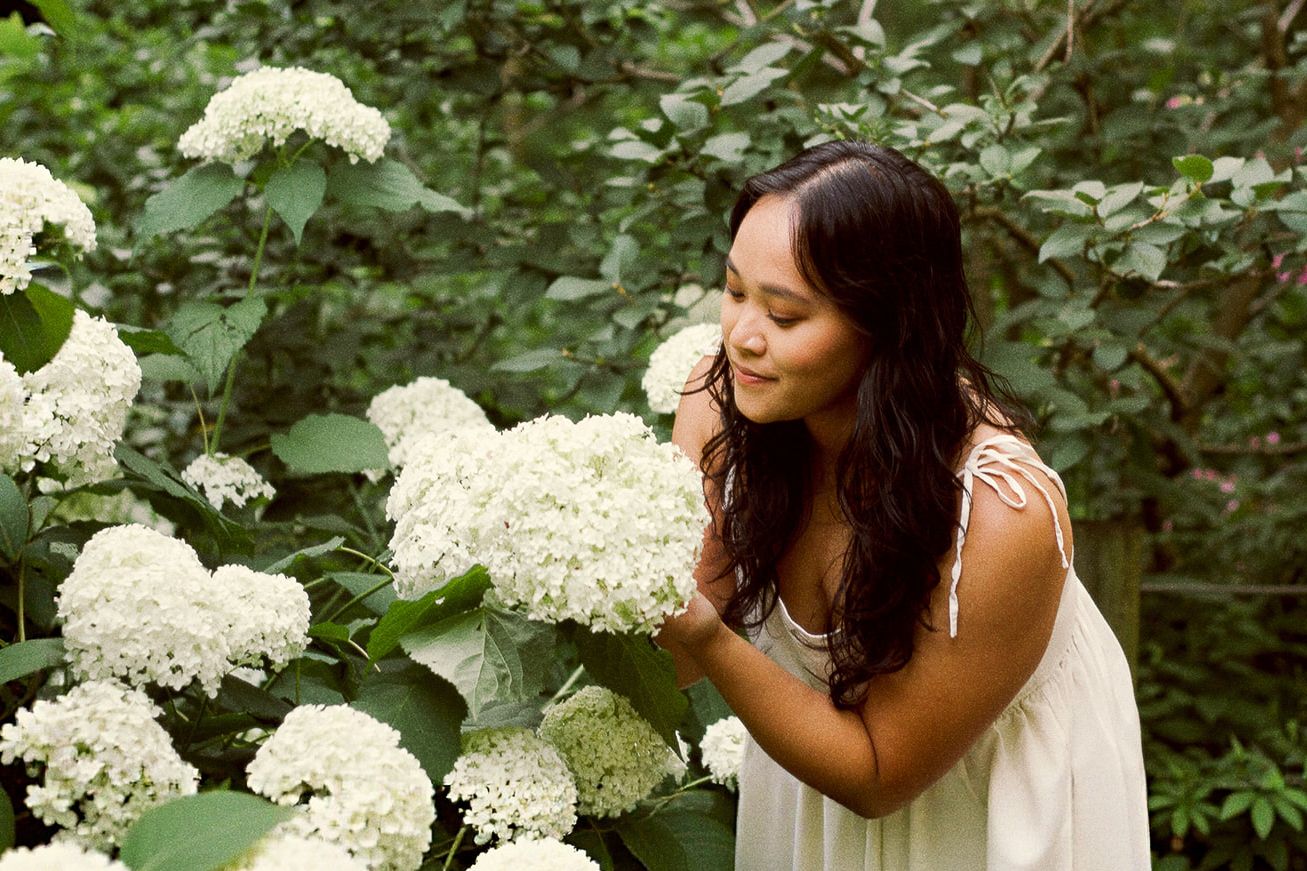 Young woman smelling white hydrangea flowers in a lush garden, wearing a white dress. DMV film inspired photographer