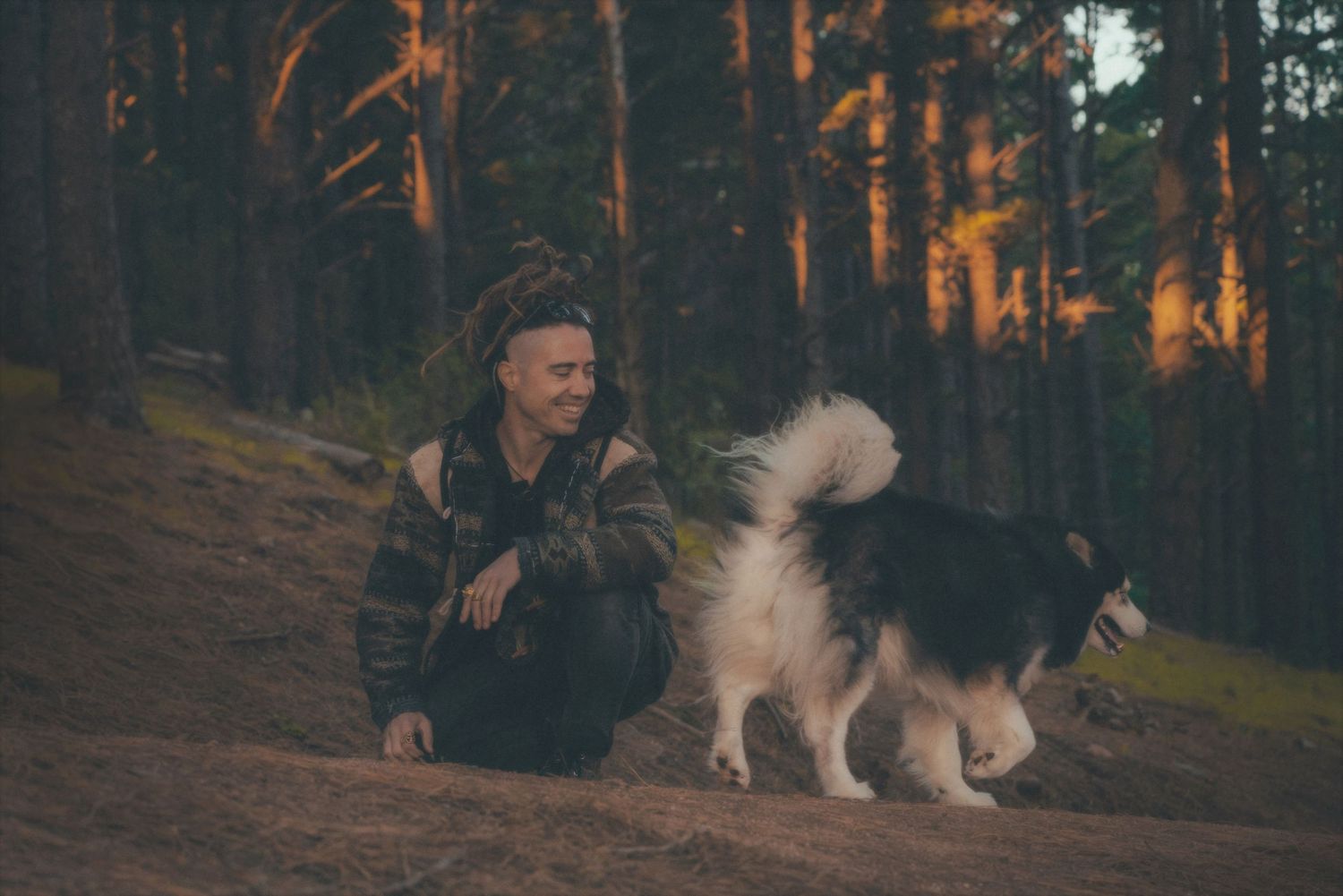 Man crouching next to his husky among the pine trees