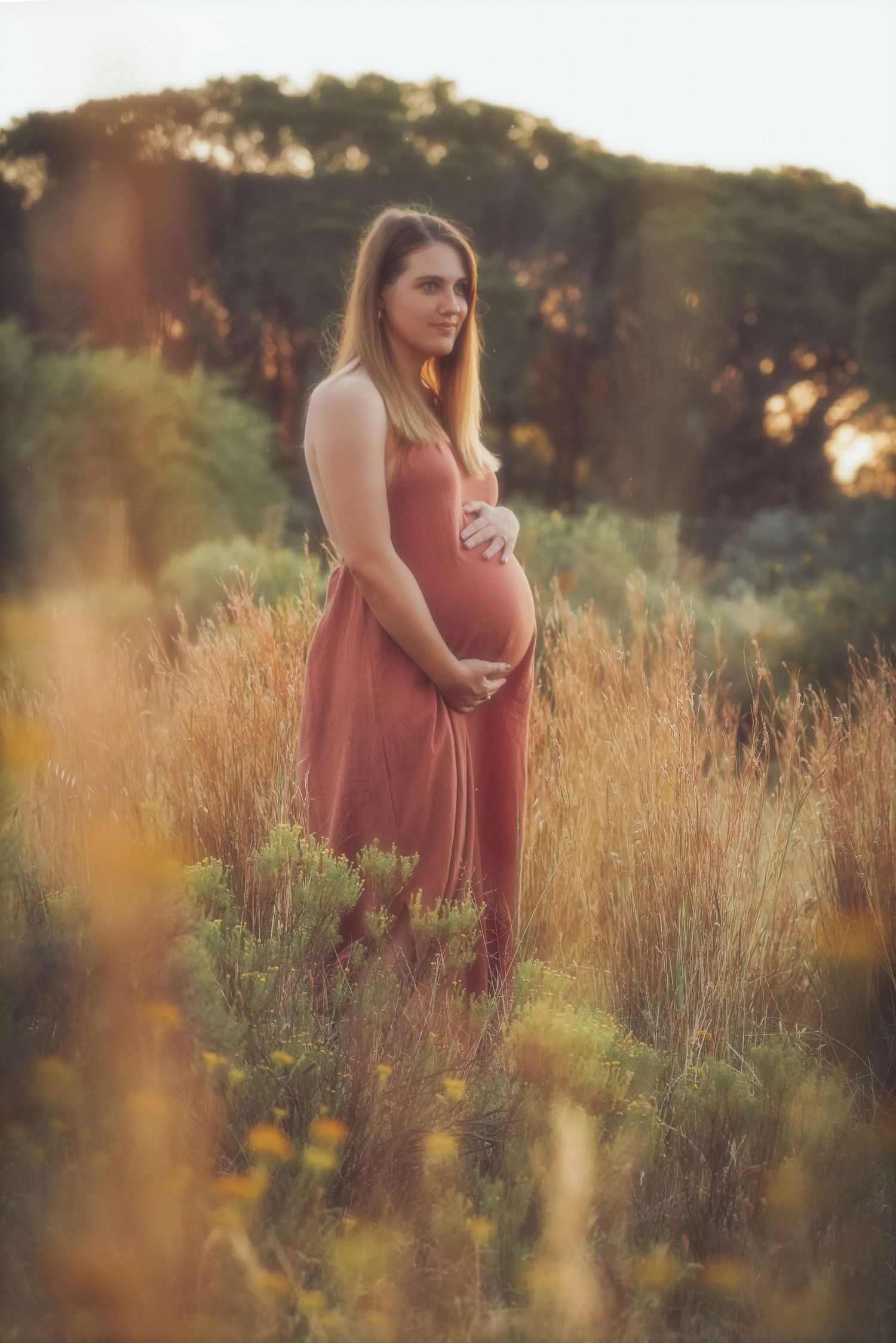 Pregnant woman in a terracotta dress standing in tall grass, hand on her bump