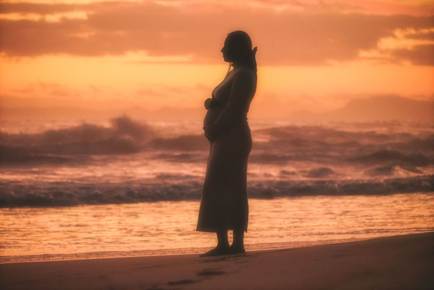 Silhouette of a pregnant woman at the water's edge against an orange sunset