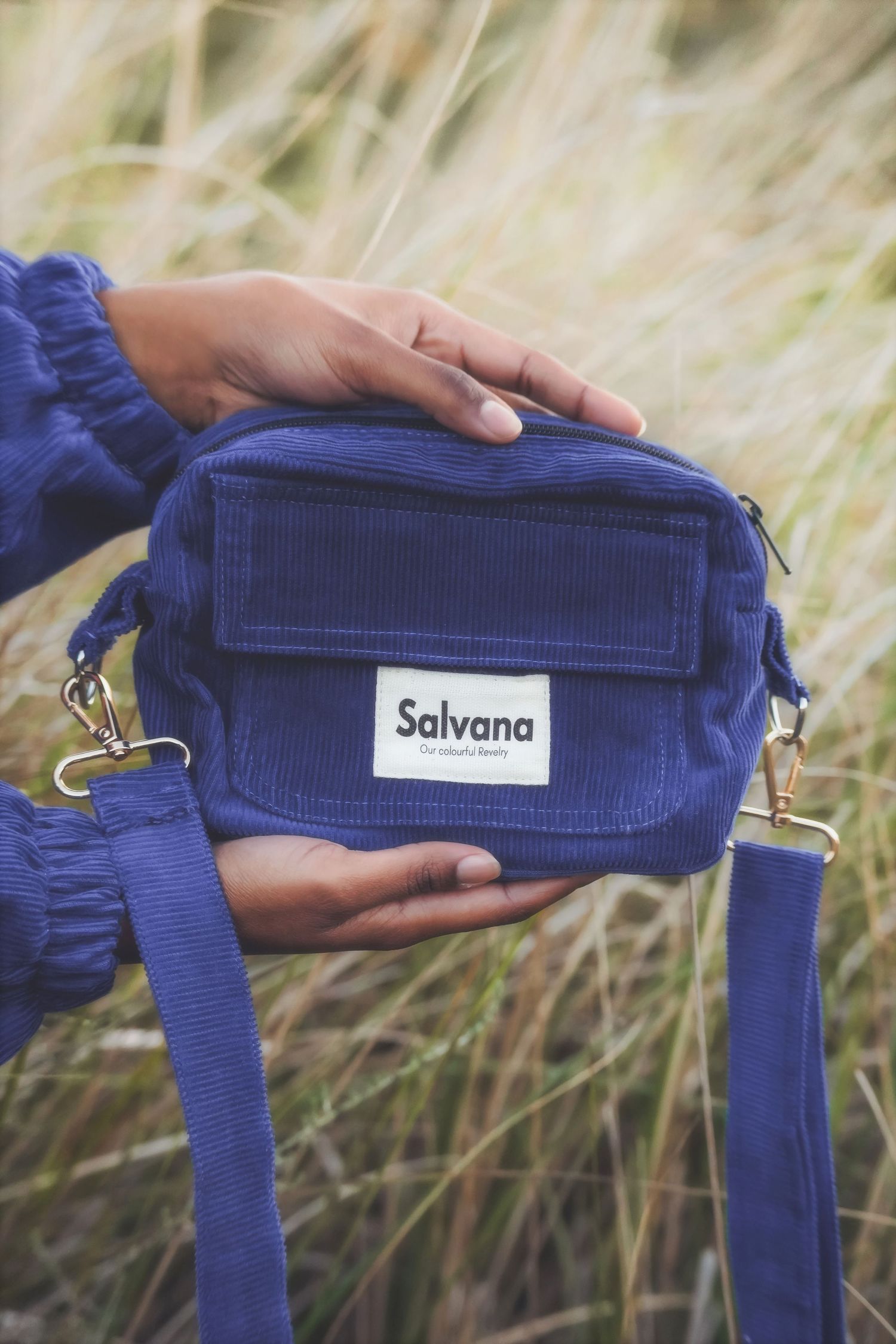 Hands holding a blue Salvana corduroy crossbody bag in tall grass